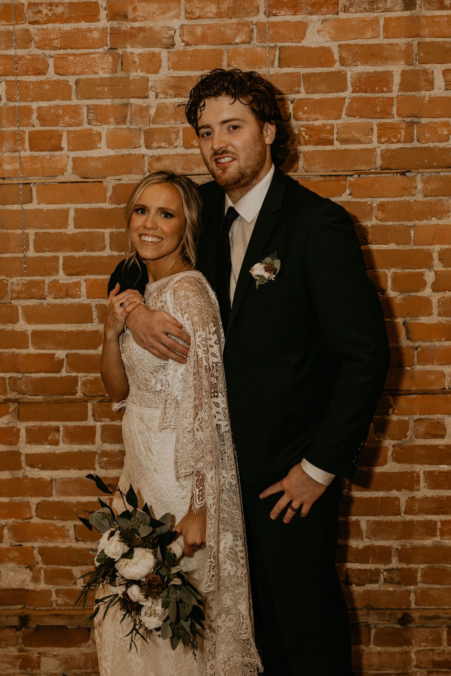 Elegant wedding portrait featuring lace dress and classic black tuxedo against rustic brick backdrop.