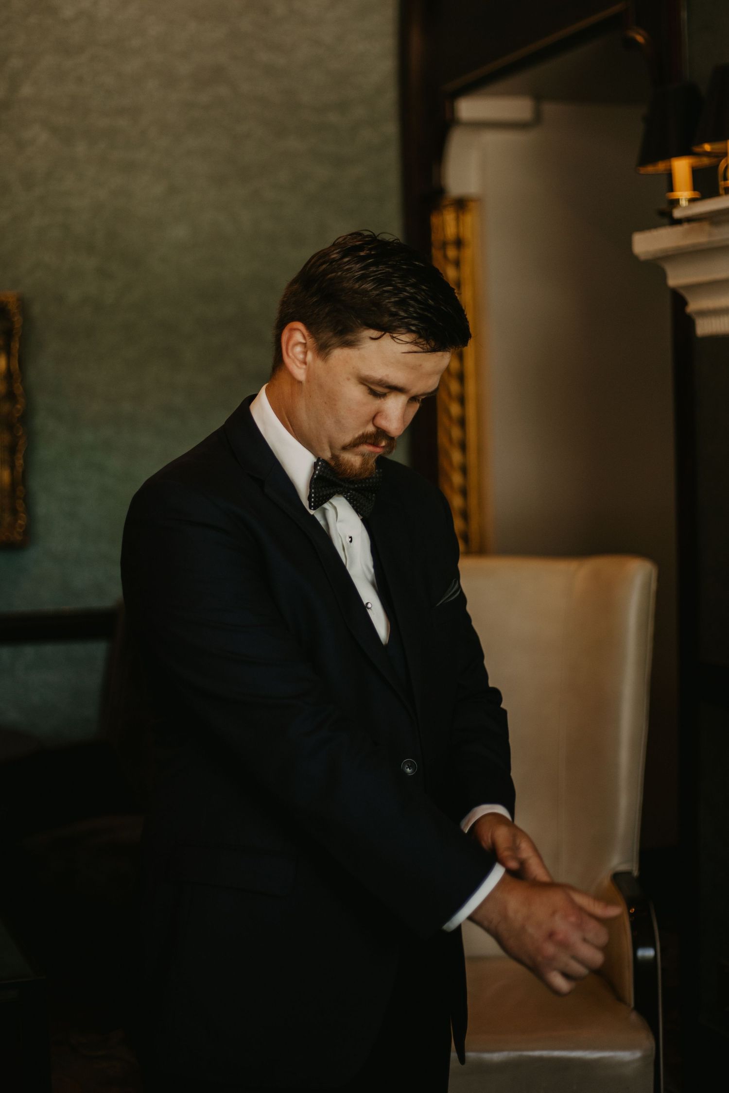 Person in formal black tuxedo adjusts cuffs in elegant room with decorative molding.
