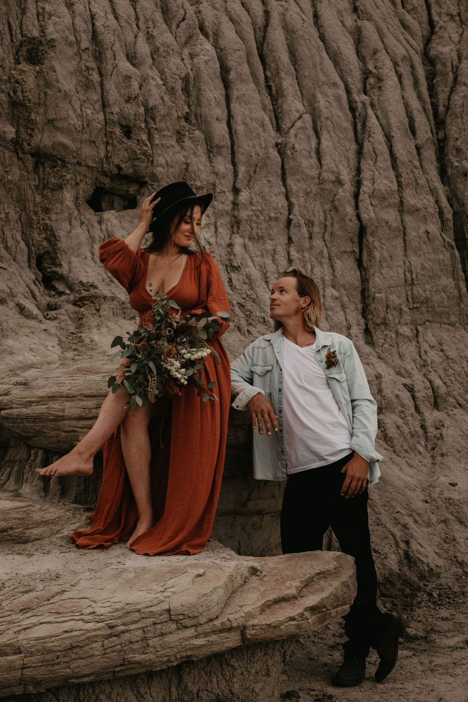 A stylish couple posing against dramatic clay cliffs with the woman wearing a flowing rust-colored dress.