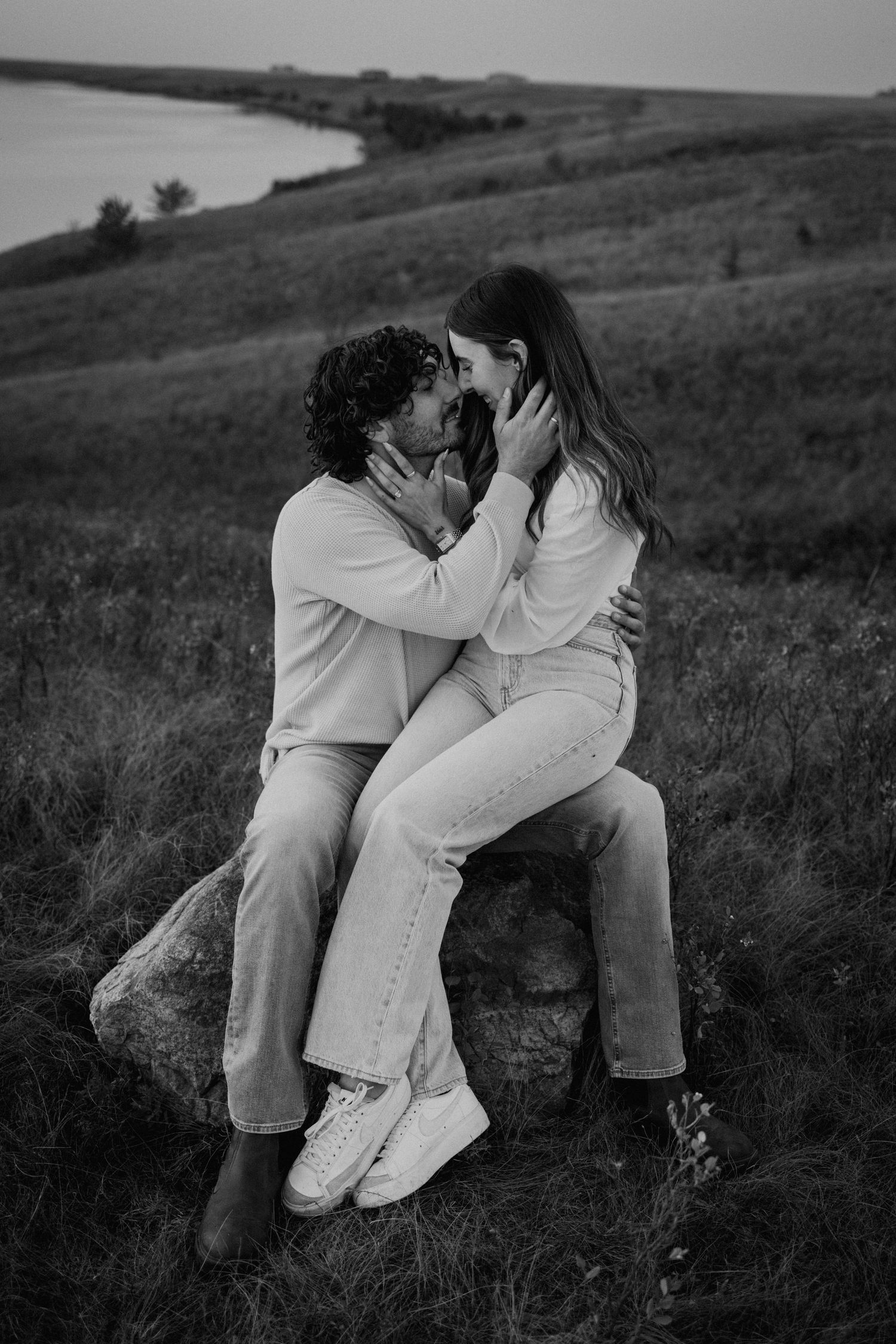 Dreamy black and white photo of couple embracing on hillside surrounded by tall grass.
