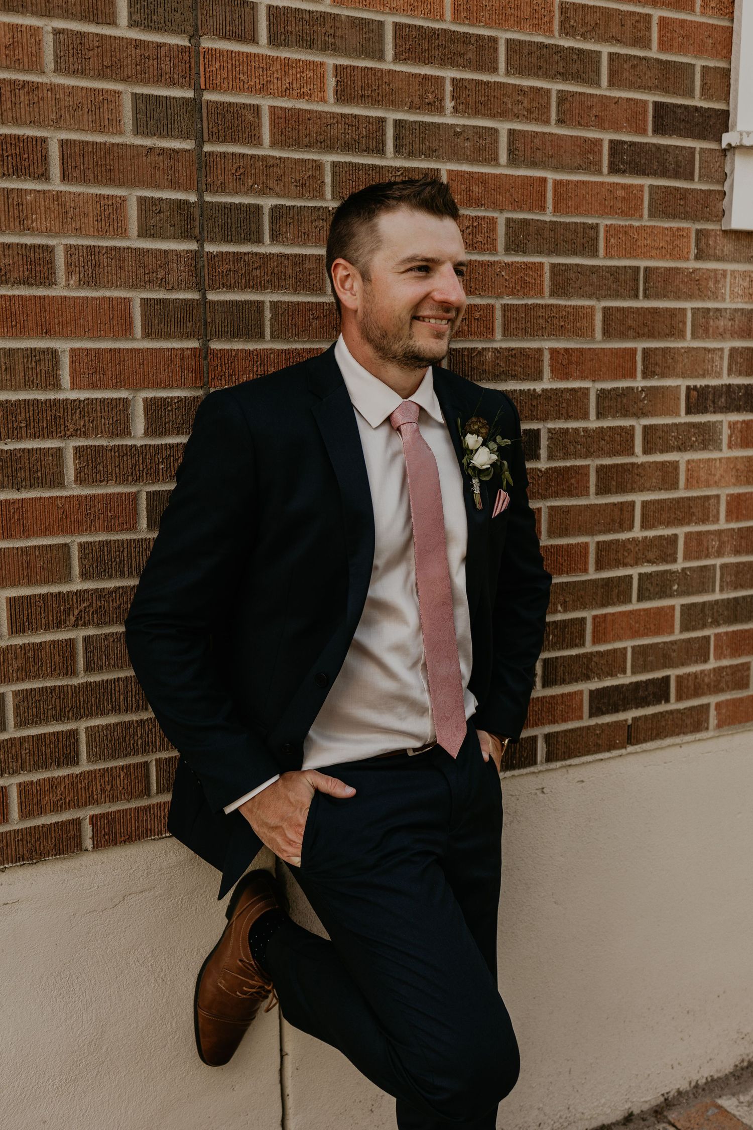 A groom in a dark suit and pink tie smiles while leaning against a brick wall.