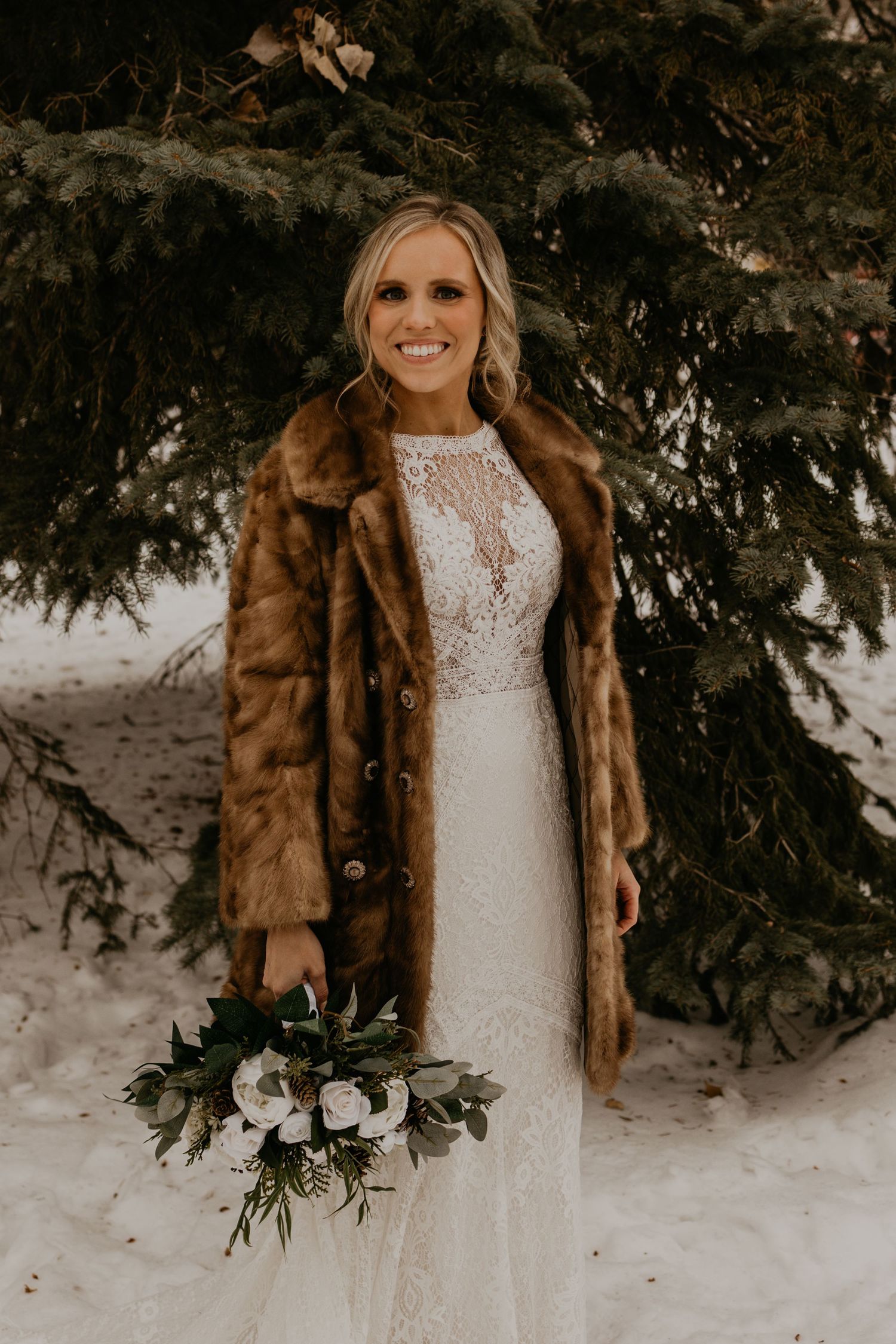 Bride in white lace dress and brown fur coat smiles while holding winter bouquet in snowy setting.