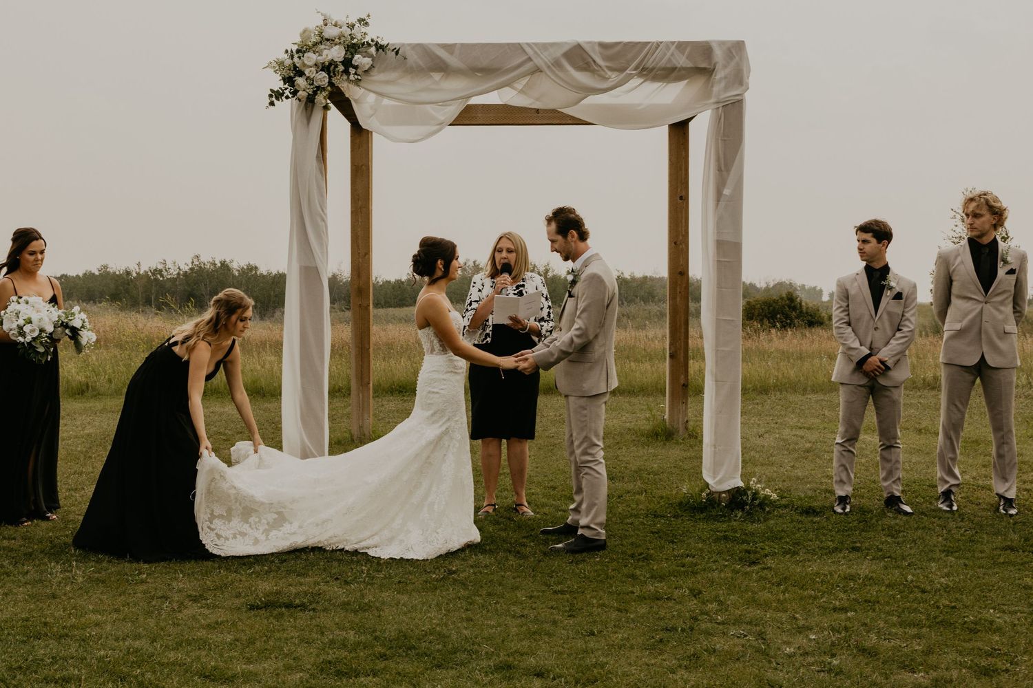 A wedding ceremony under a white canopy with attendants in beige and black watching.
