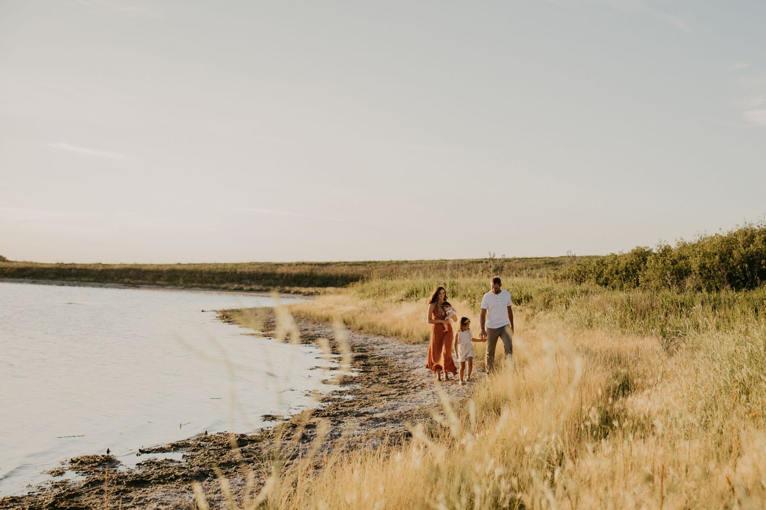 Family walking along a scenic shoreline at sunset with tall grass and water views.