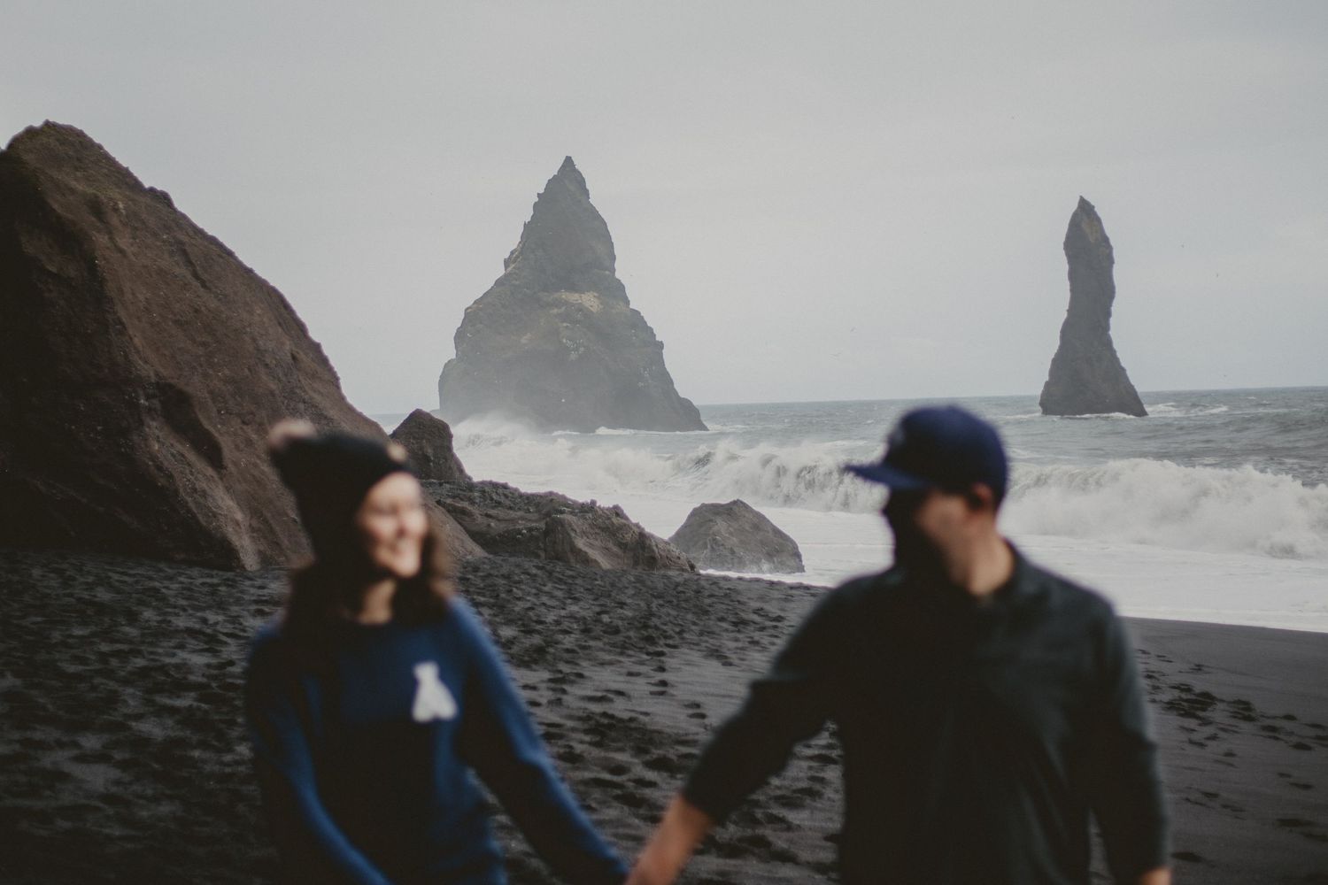 Couple walking along dramatic black sand beach with towering sea stacks in Iceland.
