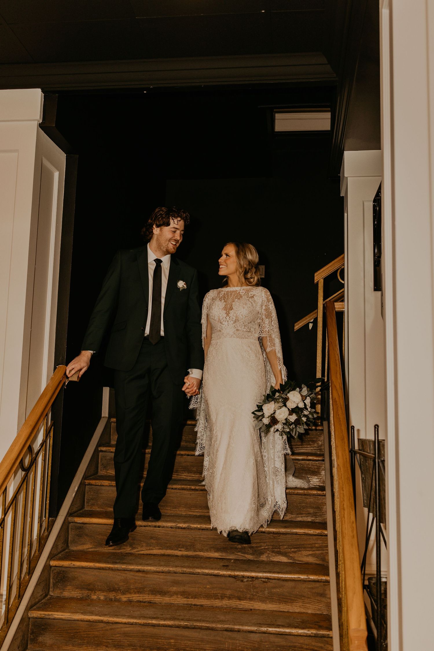A newlywed couple descends a wooden staircase together, with the bride wearing a flowing white lace dress.