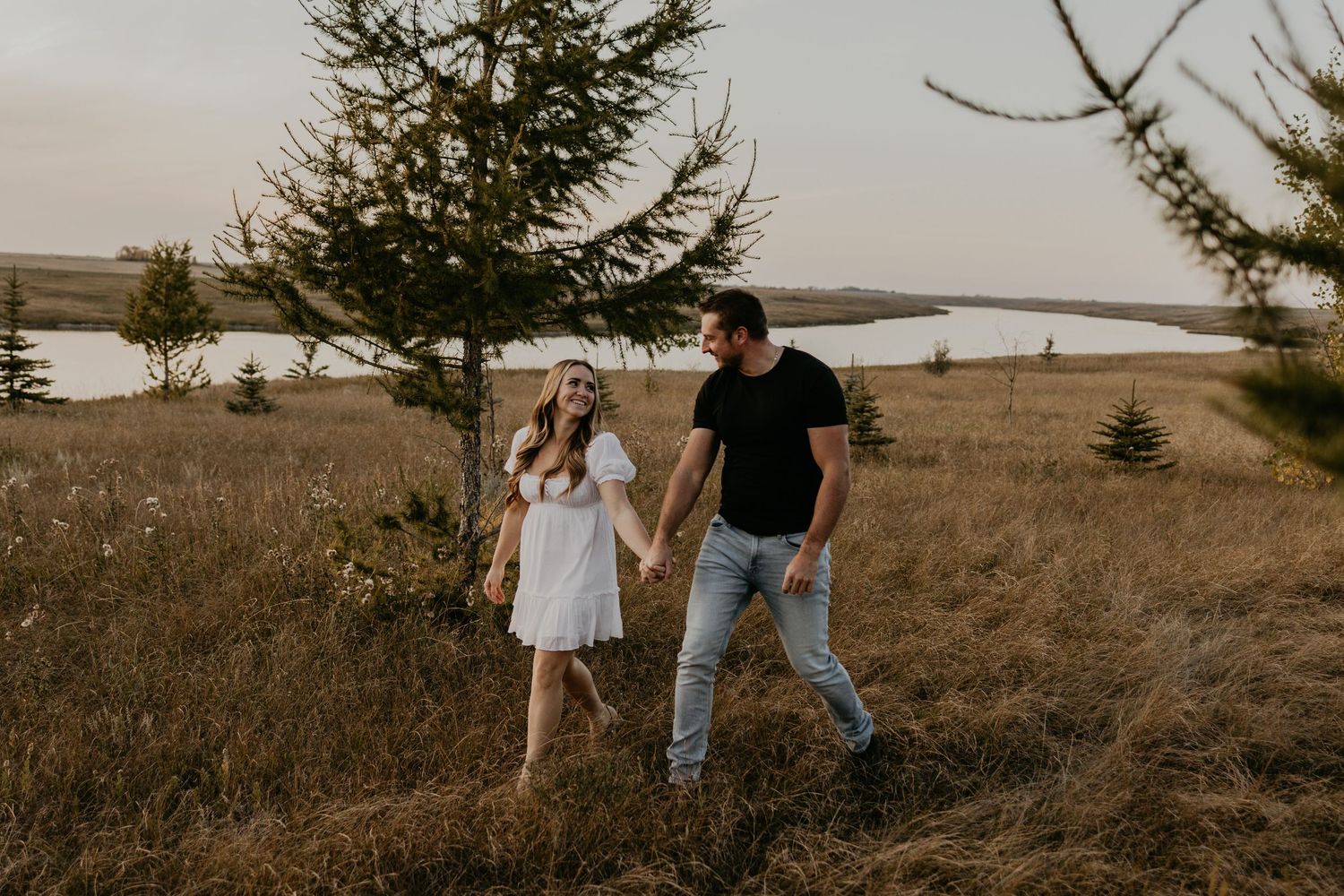 A couple walks hand in hand through a field with evergreen trees and water in the background during sunset.