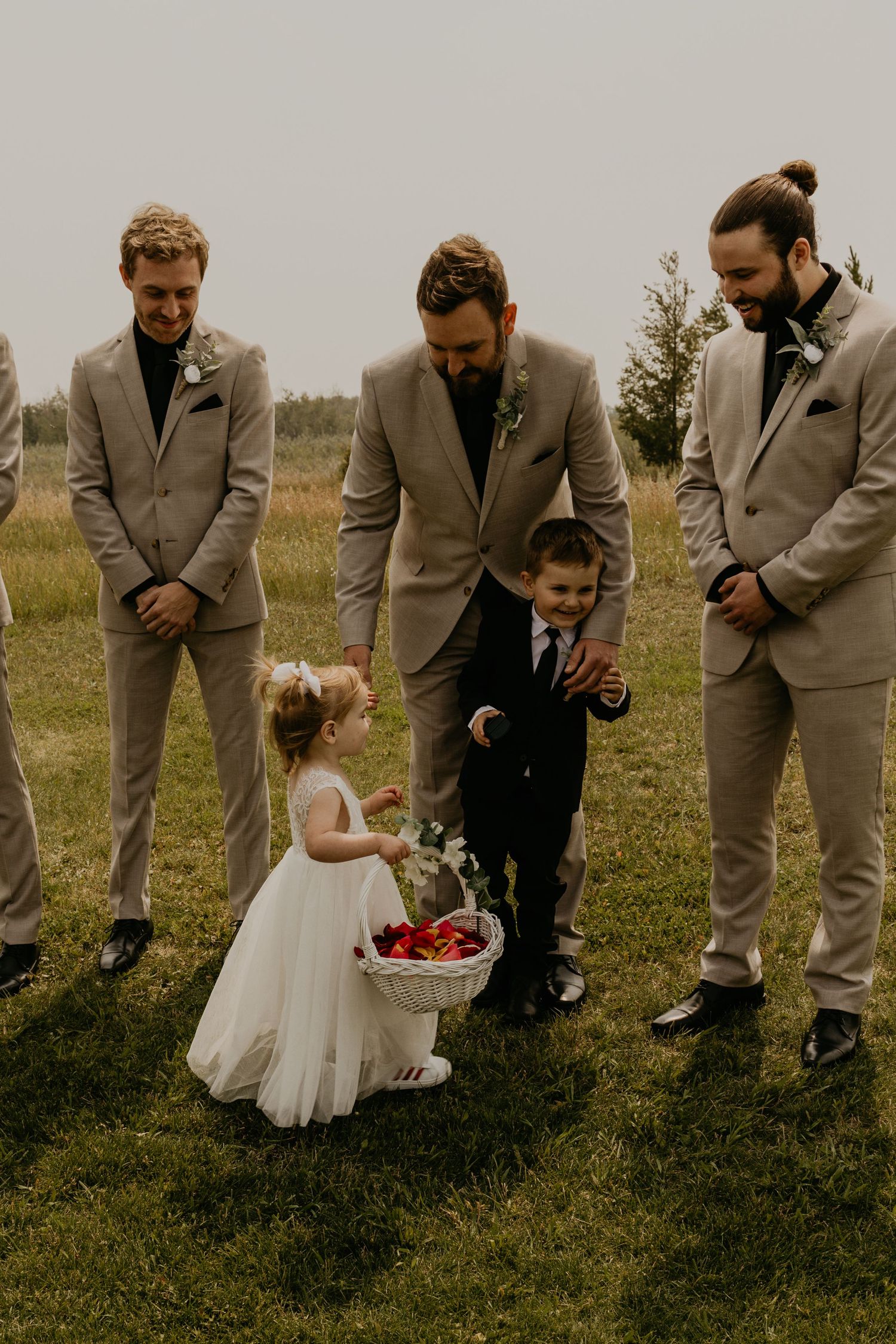 Groomsmen in beige suits watch as a flower girl with a basket interacts during an outdoor wedding ceremony.