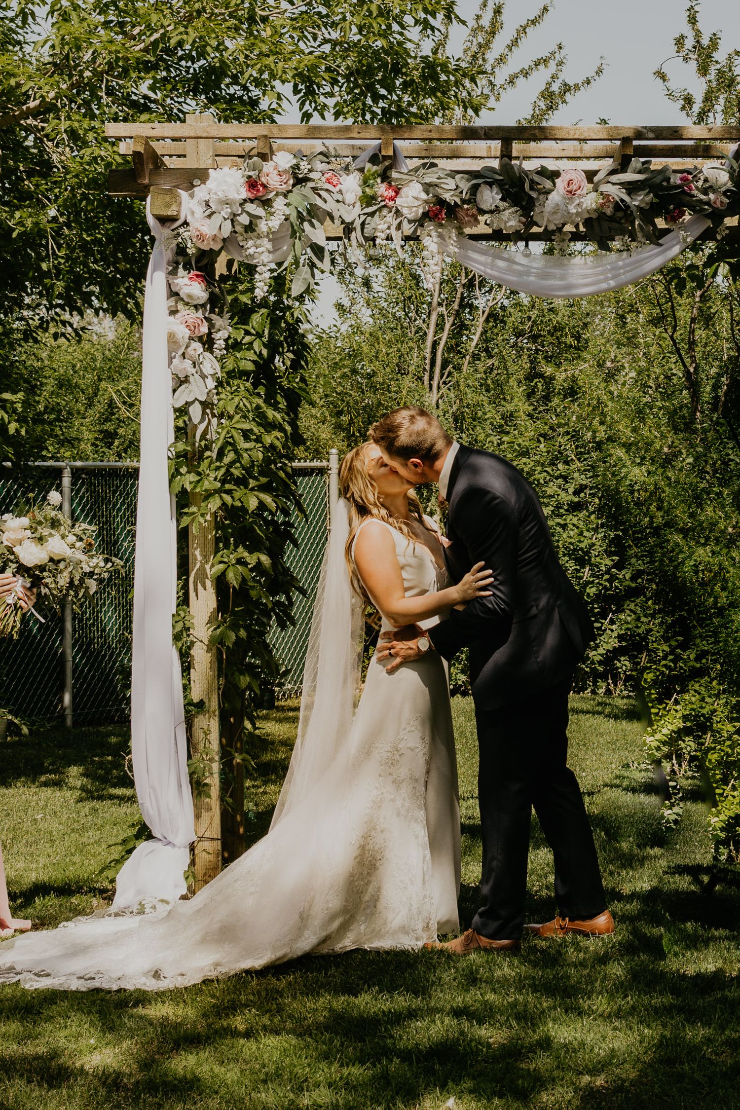 A romantic wedding ceremony under a floral arch decorated with white fabric and flowers in a sunlit garden setting.