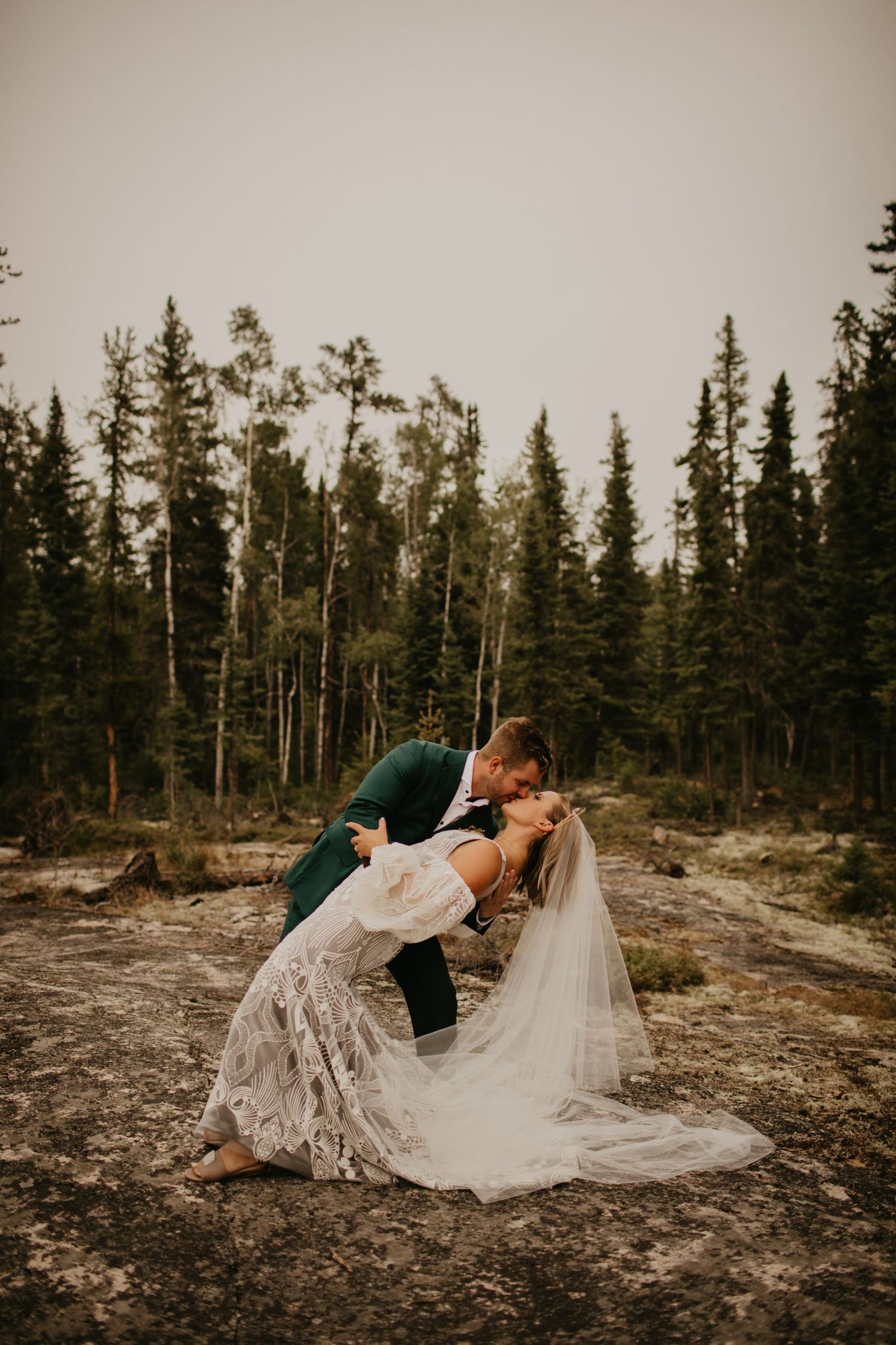Romantic wedding pose on a rocky terrain with evergreen forest backdrop.