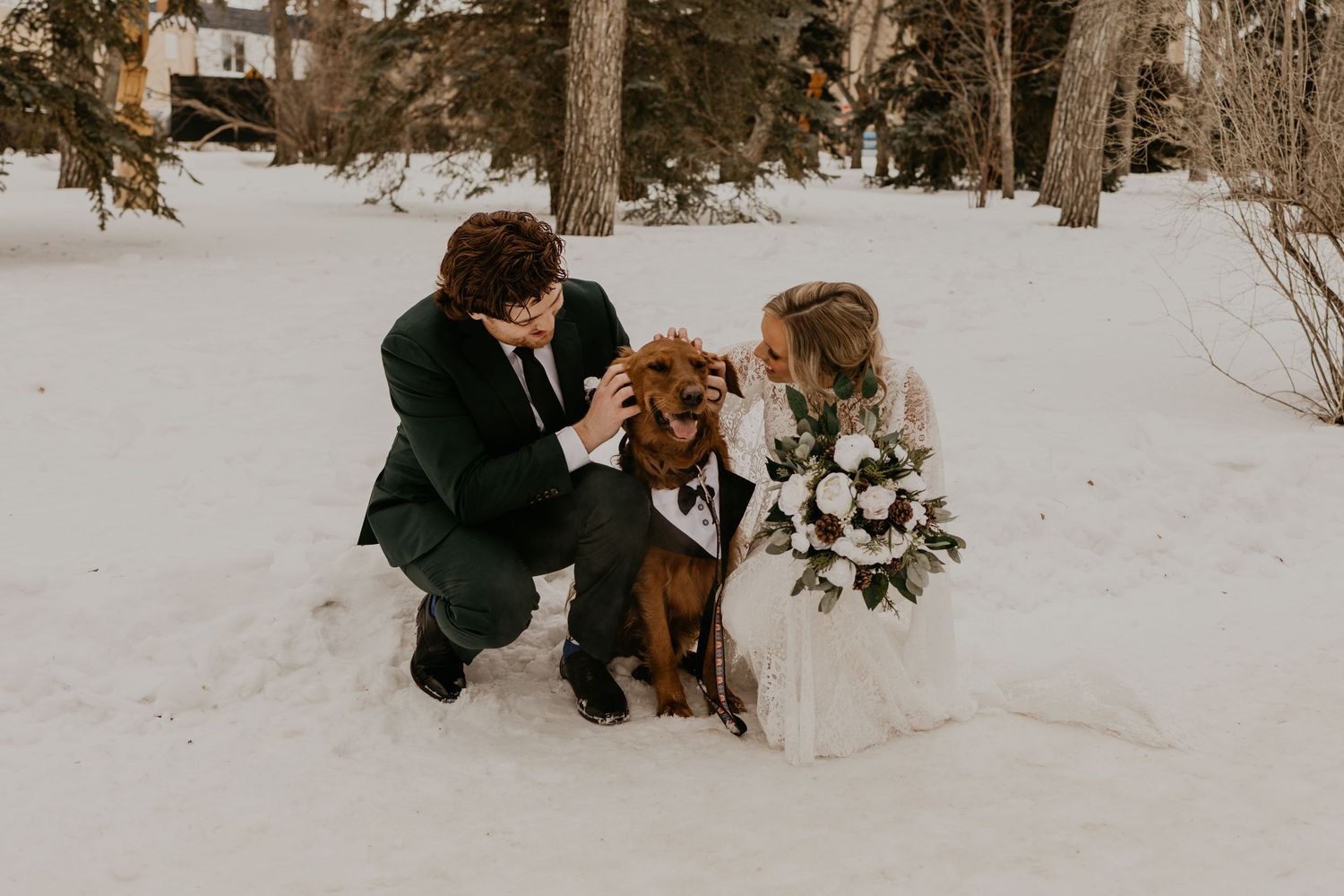 Couple kneels with their dressed-up dog in snowy winter scene during wedding photoshoot.