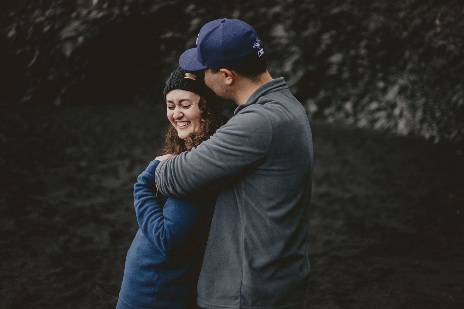 A couple shares laughter and embraces against a dramatic dark rock wall background.