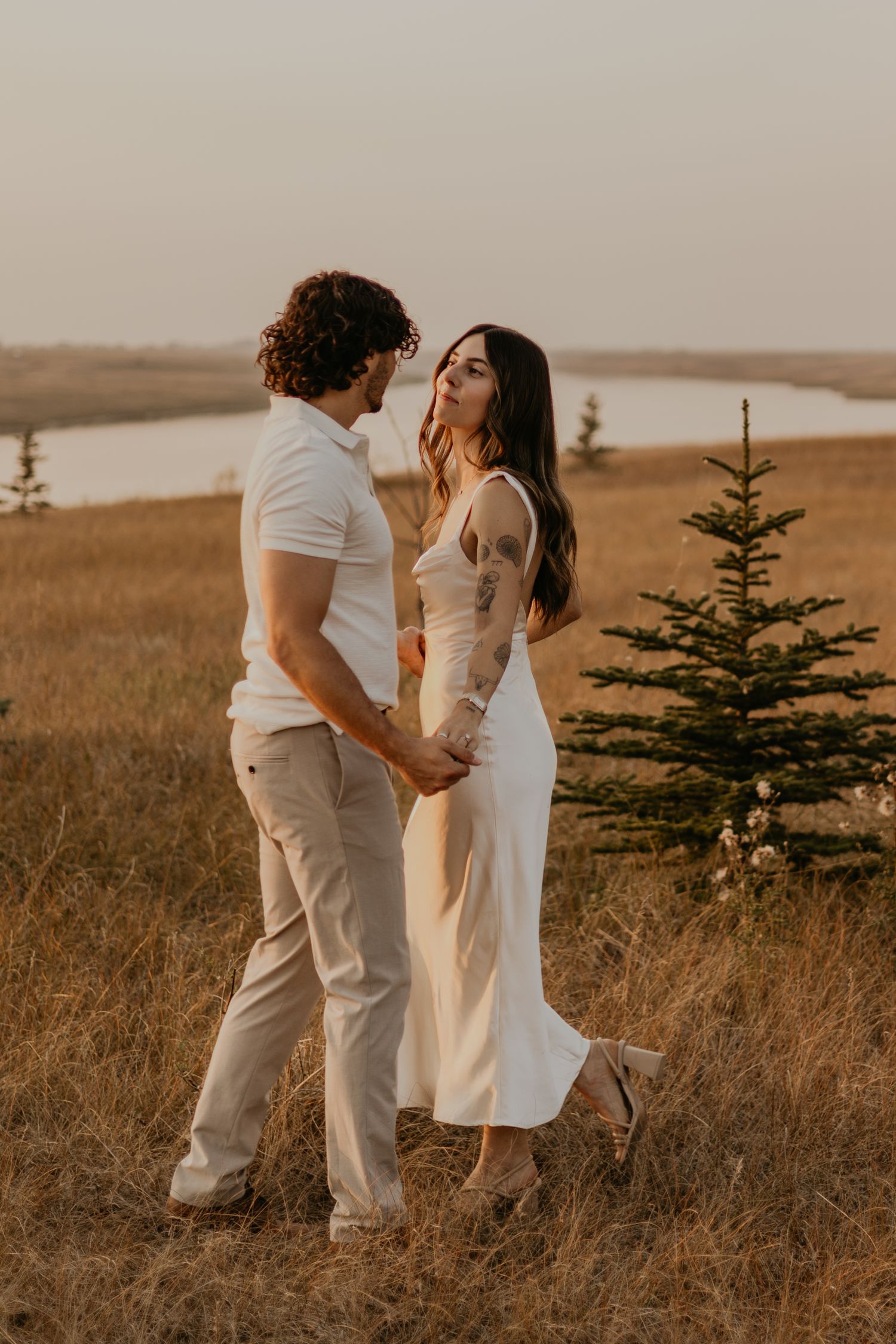 Couple in white and beige outfits share intimate moment on hilltop at golden hour.