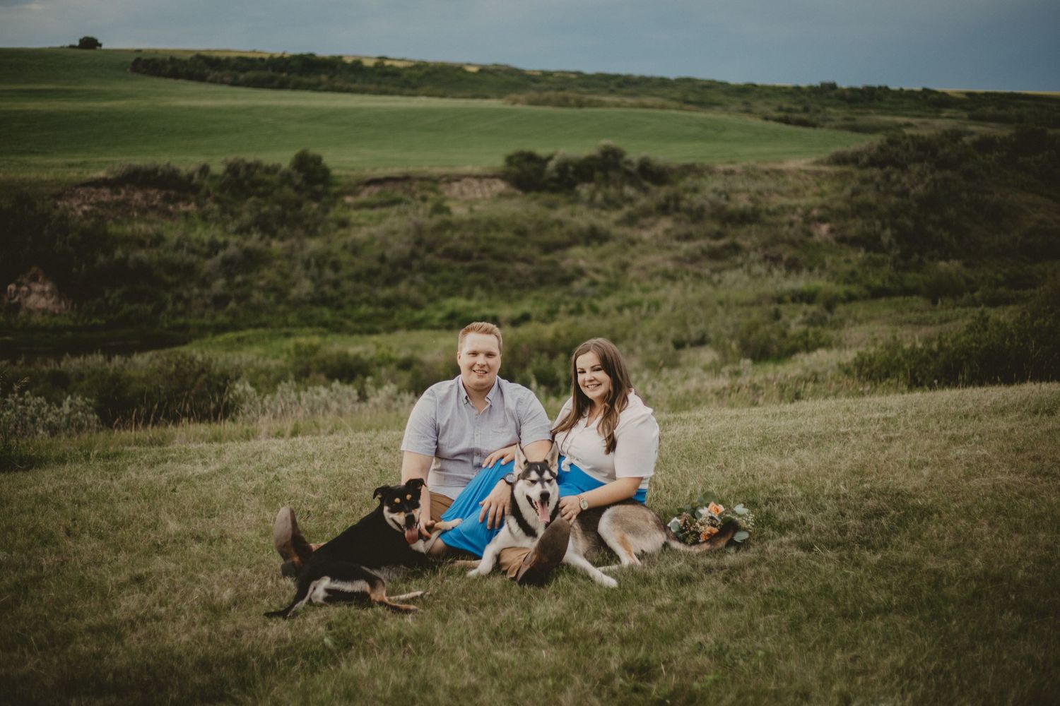 A couple sits with their dogs on a grassy hillside during a sunny afternoon.
