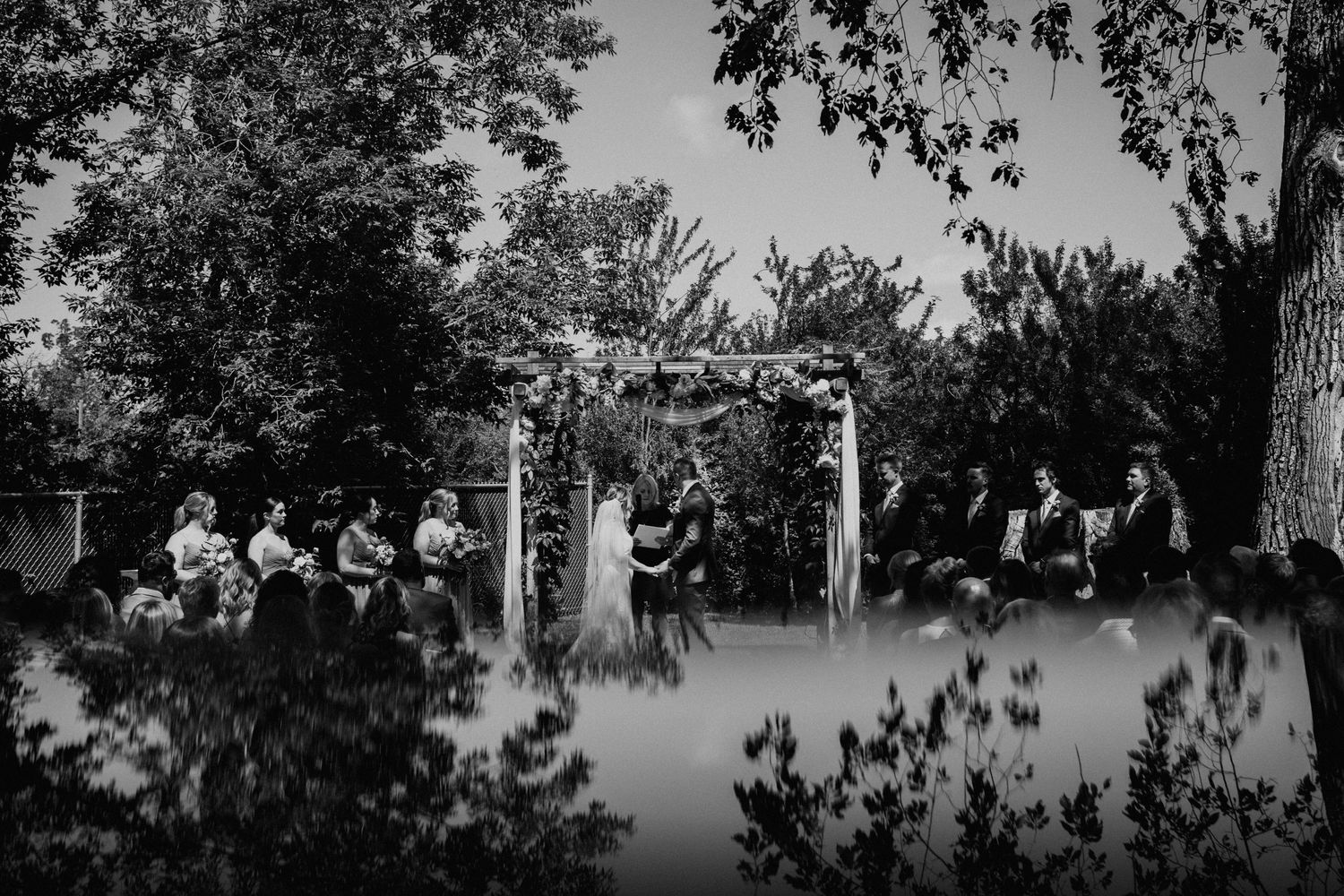 Black and white photo of an outdoor wedding ceremony under an arch with guests seated on either side.