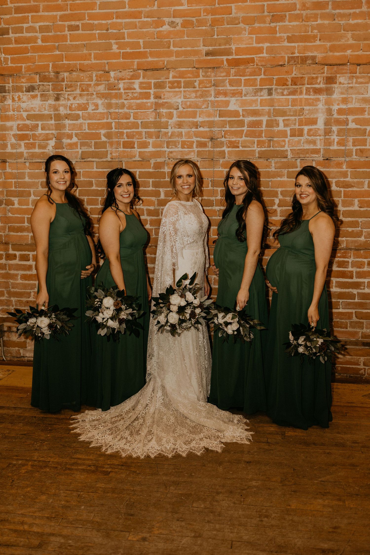 Bridal party photo showing bridesmaids in emerald green gowns with white floral bouquets against brick backdrop.