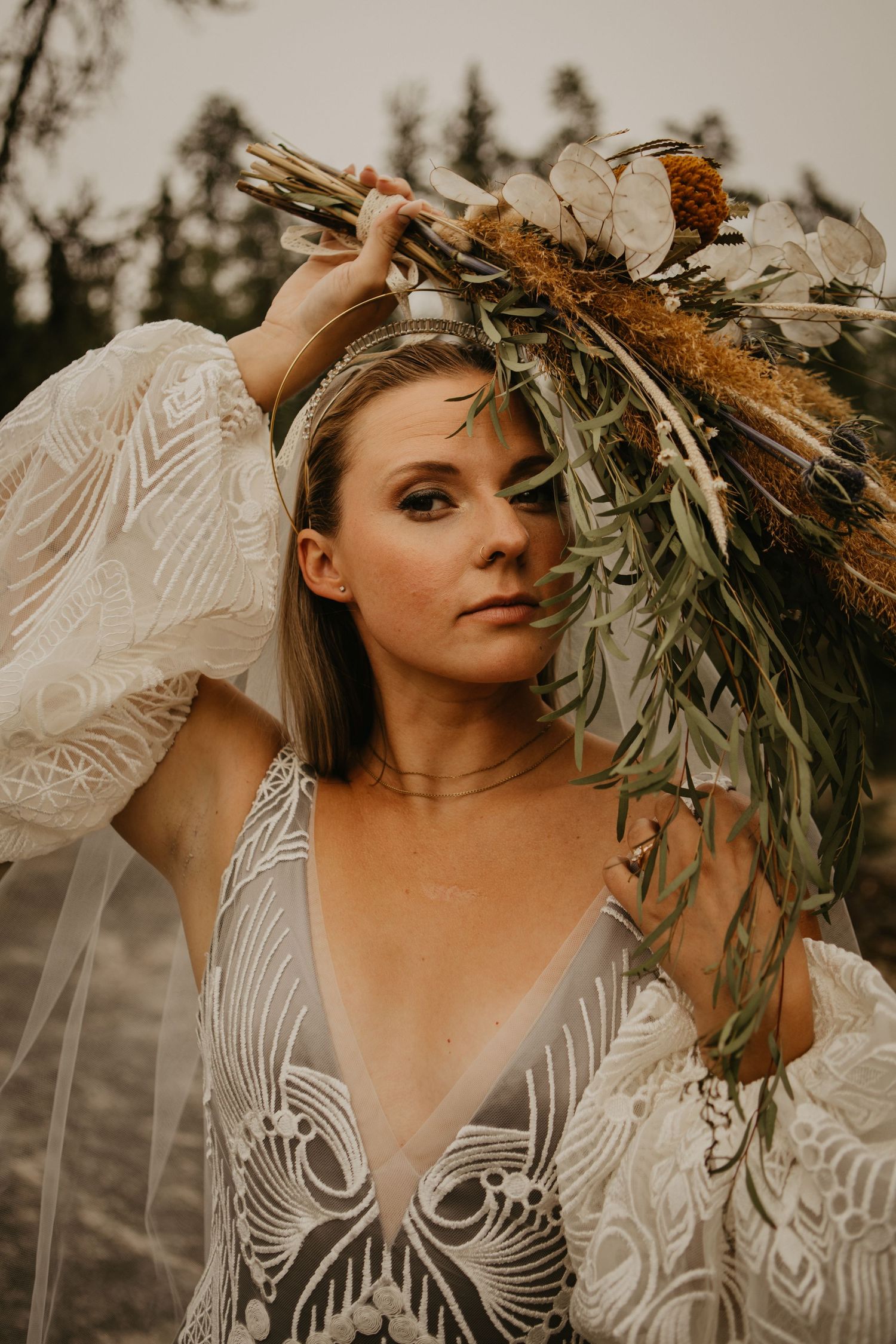 Bride holding dried floral bouquet in detailed lace wedding dress against forest backdrop.