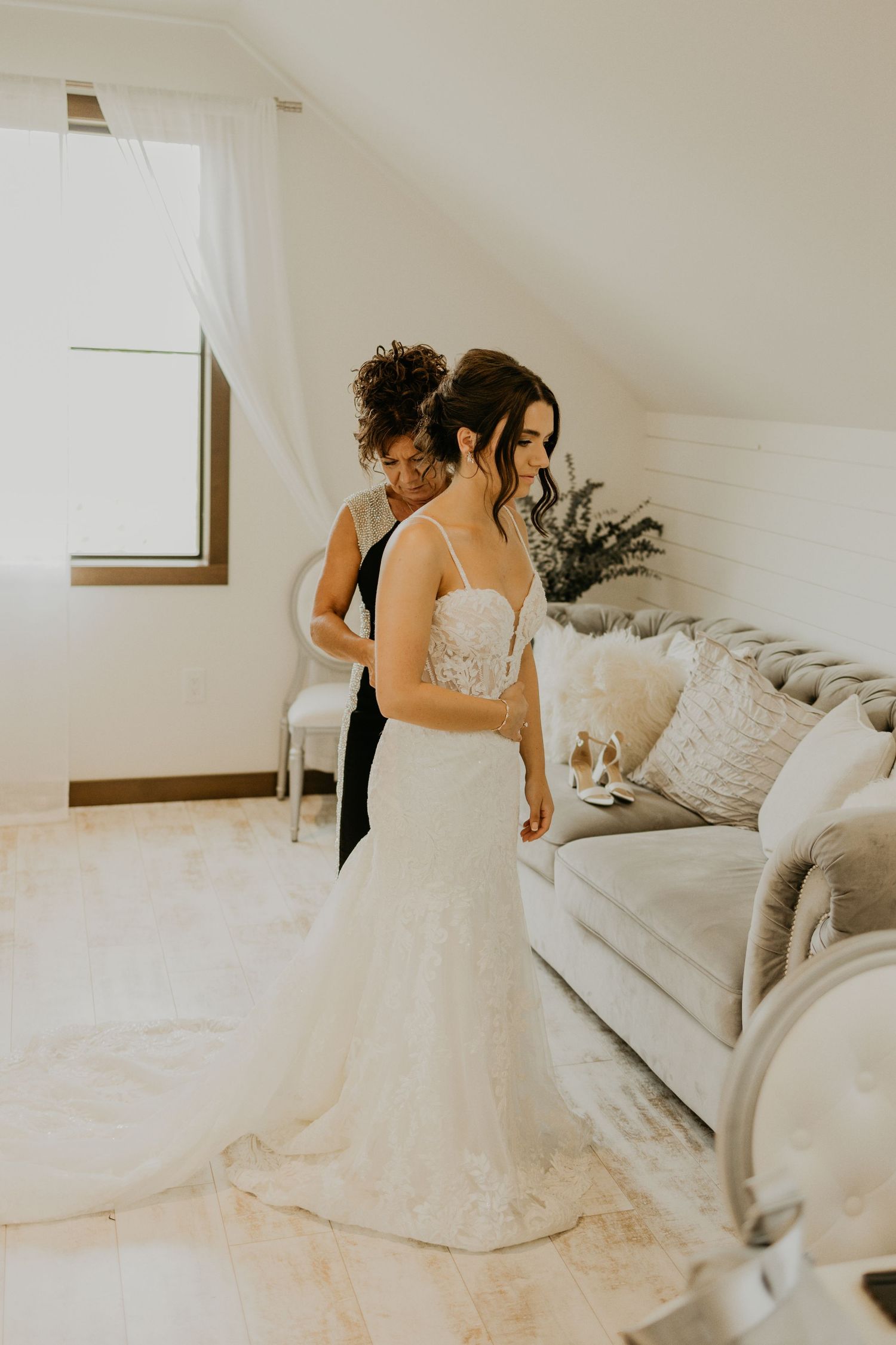 A beautiful lace wedding dress being adjusted in a sunlit room with white decor.