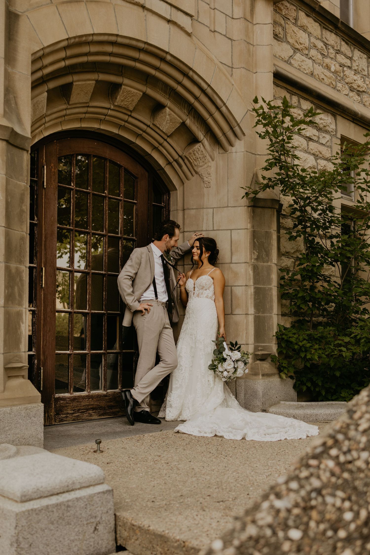 Couple in wedding attire posing against stone archway of historic building.