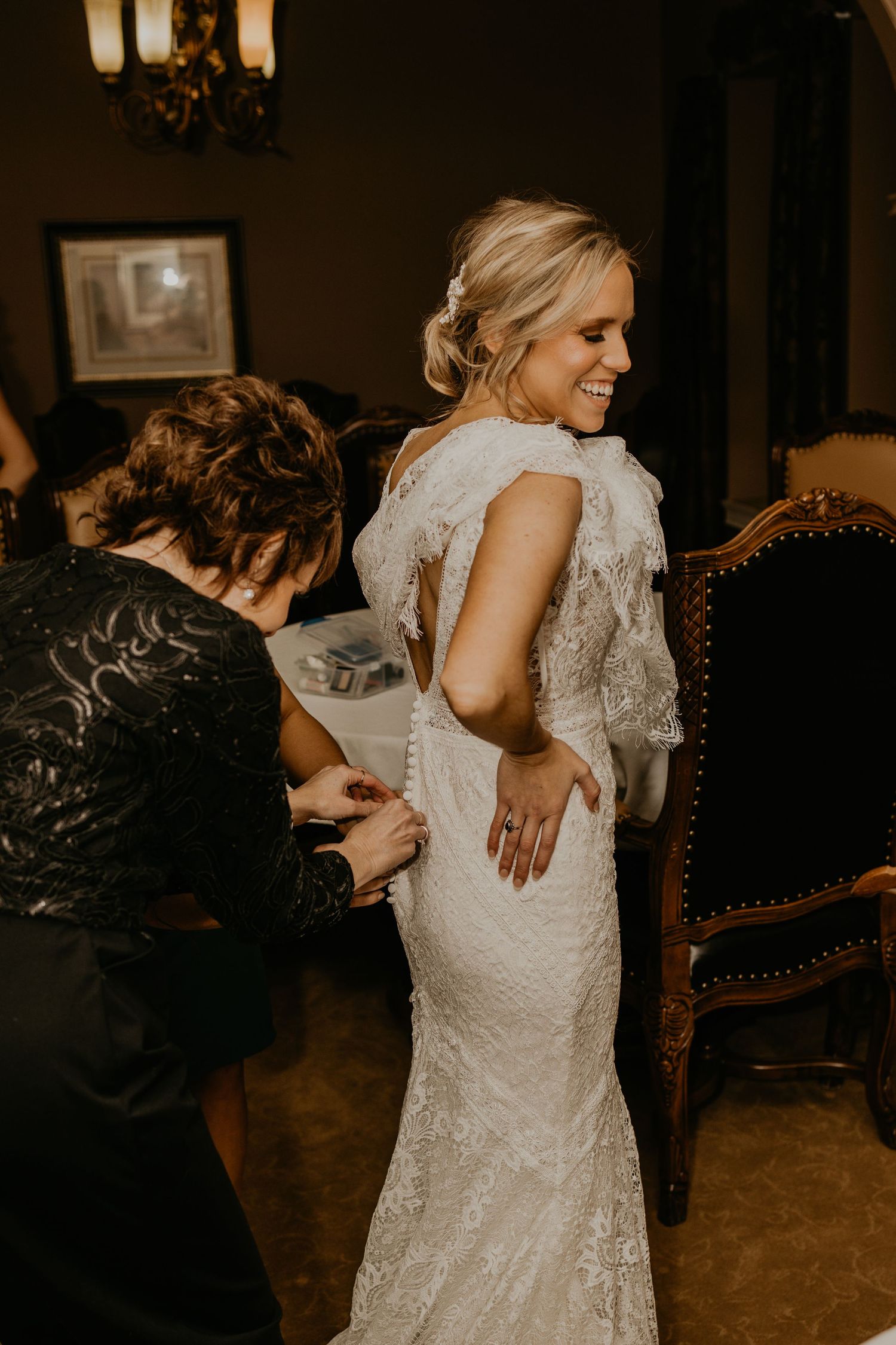 A bride getting help with her fitted lace wedding dress in a dimly lit room.