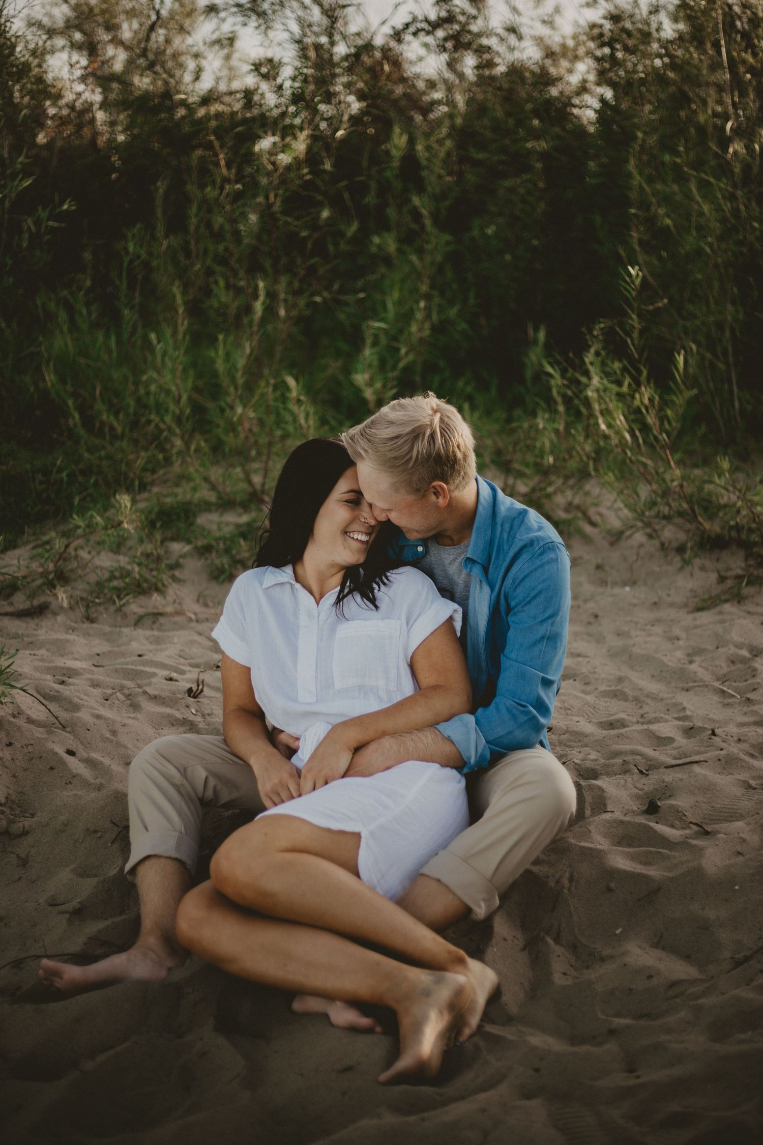 Romantic couple sitting close together on sandy beach surrounded by coastal vegetation.
