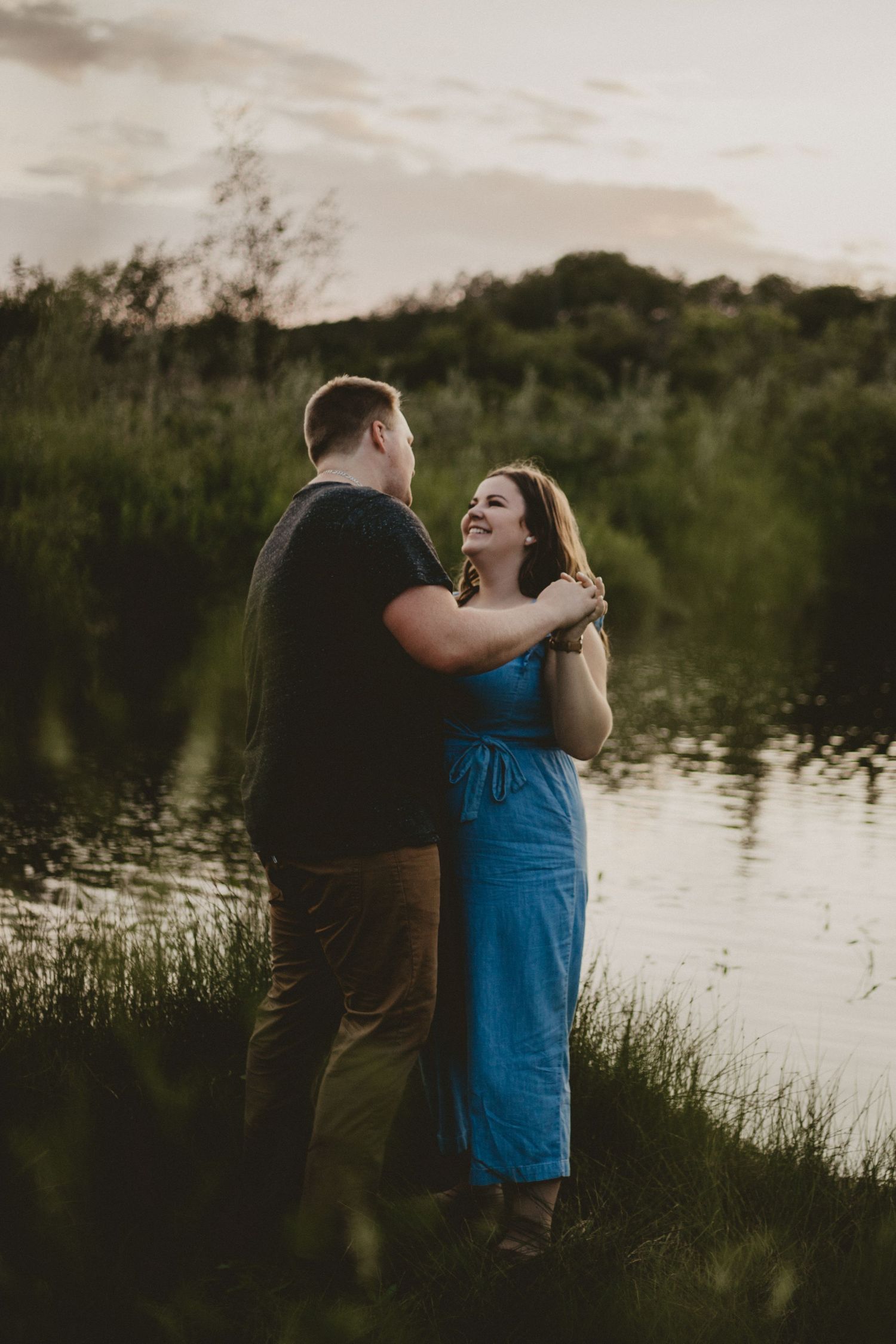 Couple embracing by a serene lake at dusk surrounded by lush greenery.