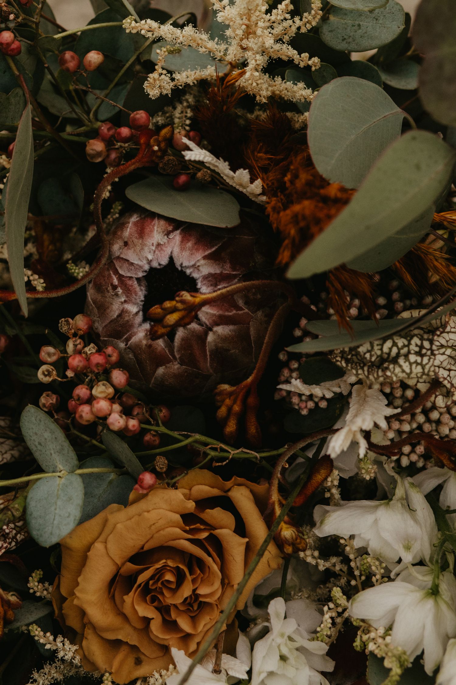 A moody floral arrangement with a golden rose, white flowers, berries, and eucalyptus leaves in dark vintage tones.