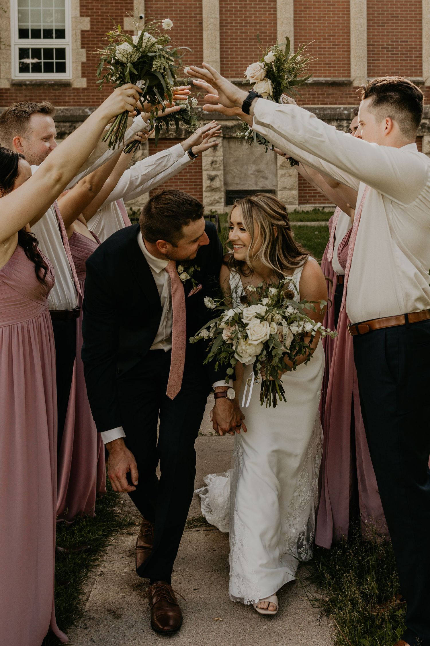Wedding party celebrates with raised bouquets as couple shares intimate moment beneath archway.