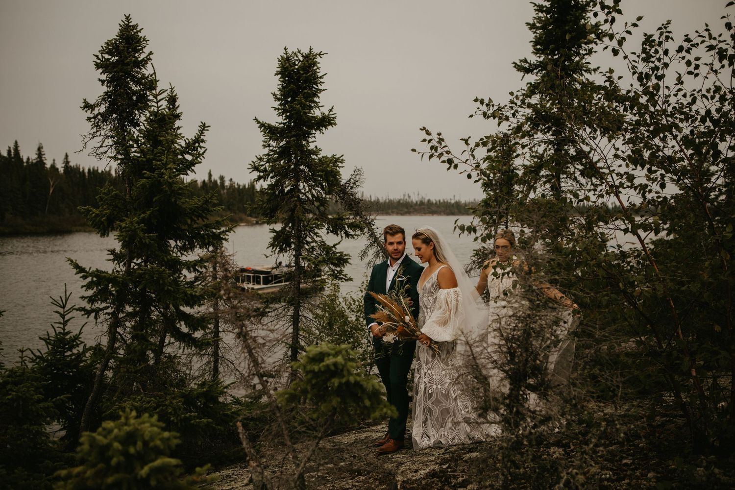 Wedding party standing on rocky overlook with lake and forest backdrop.