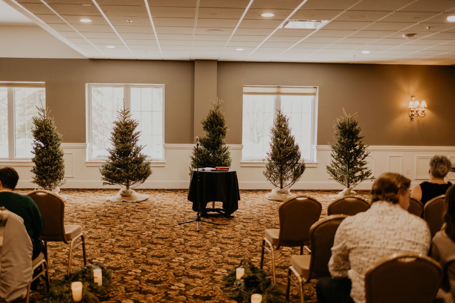 Small evergreen trees line a ceremony space with candle-lit aisle in a carpeted room.