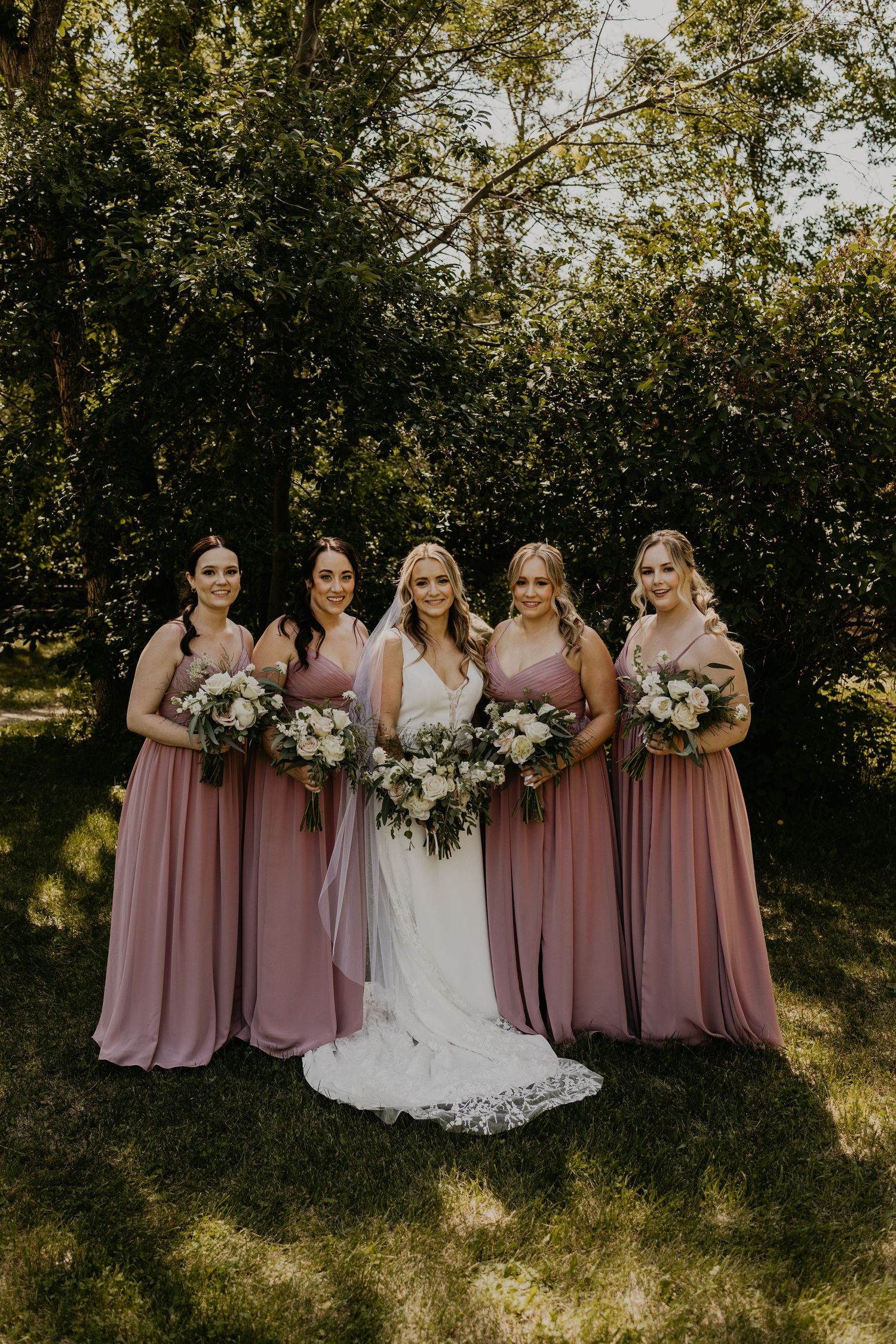 Bridal party poses with dusty rose bridesmaid dresses and white floral bouquets against woodland backdrop.