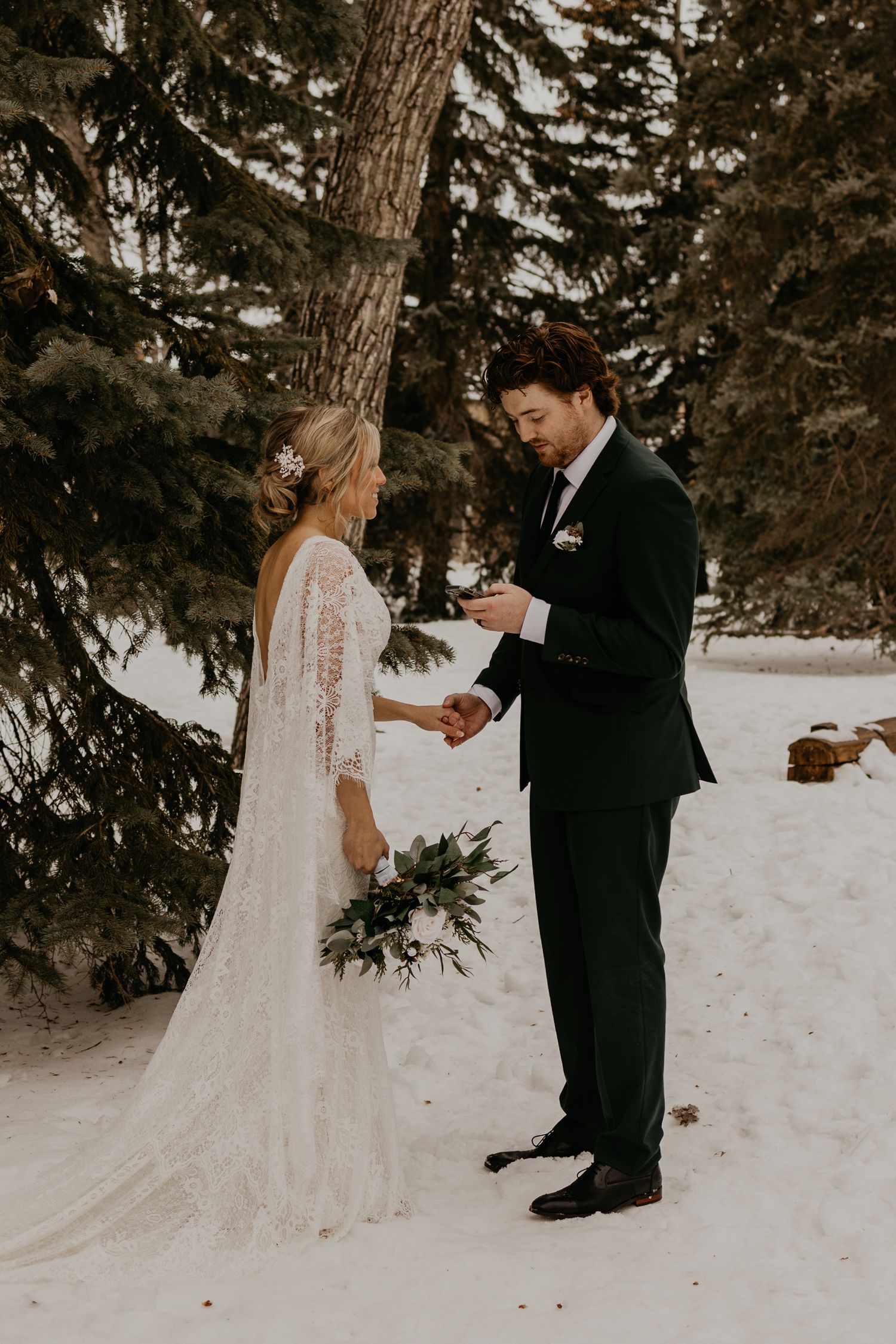 Winter wedding ceremony moment with couple reading vows in snow under pine trees.