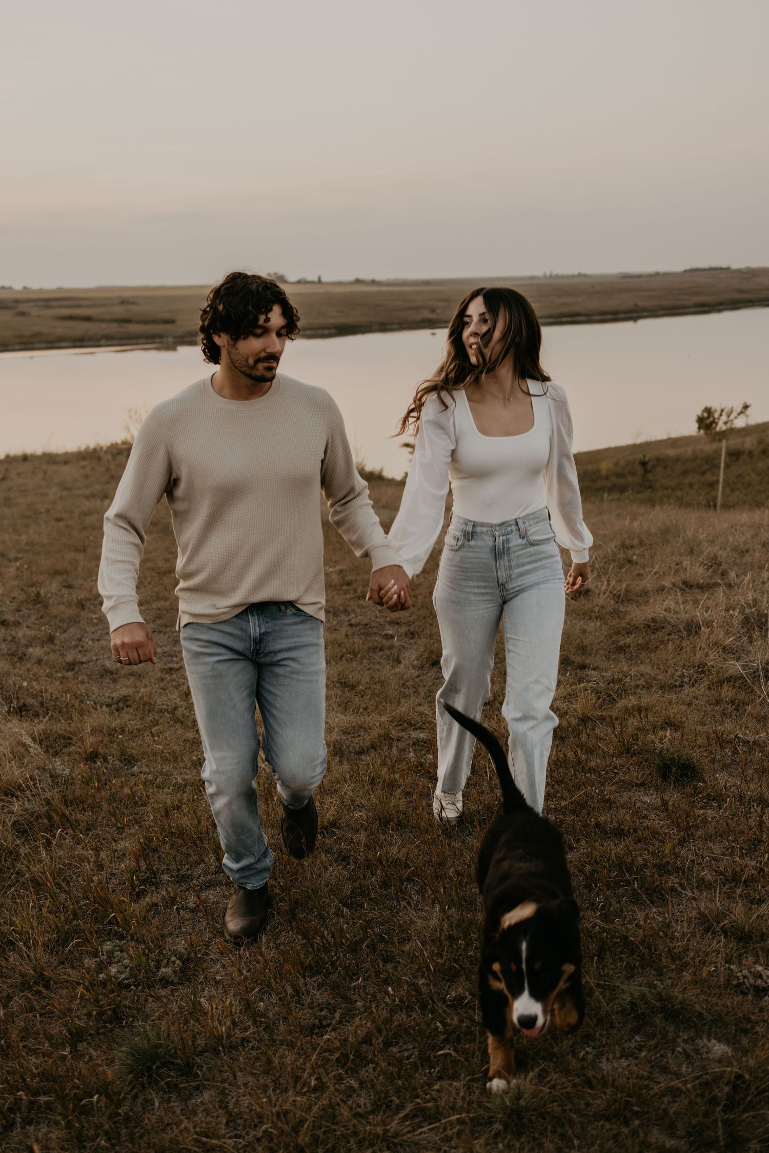 A couple walking together on a grassy hillside with their black and white border collie dog at sunset.