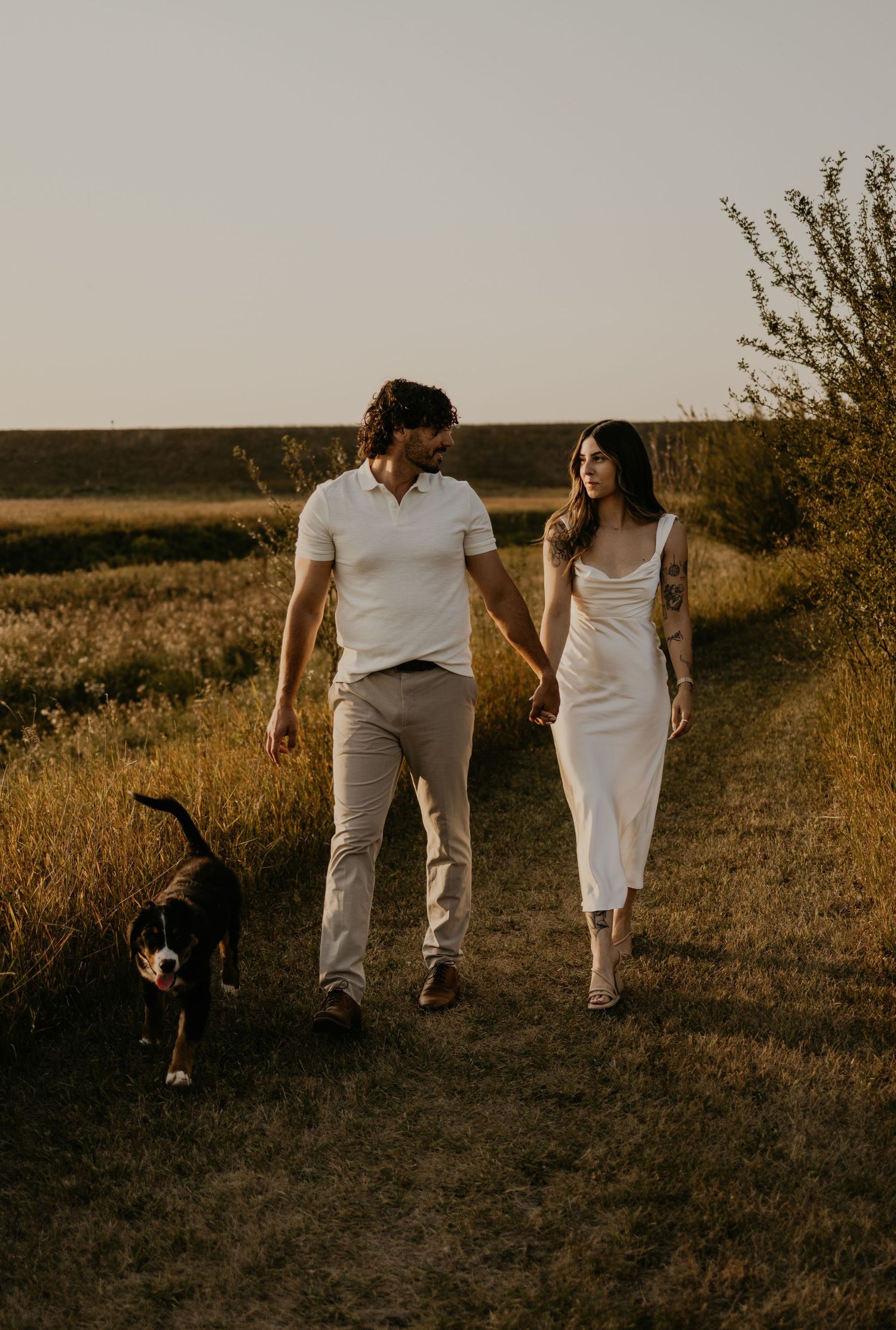 Couple in white and beige outfits walk with their dog through golden autumn fields at sunset.