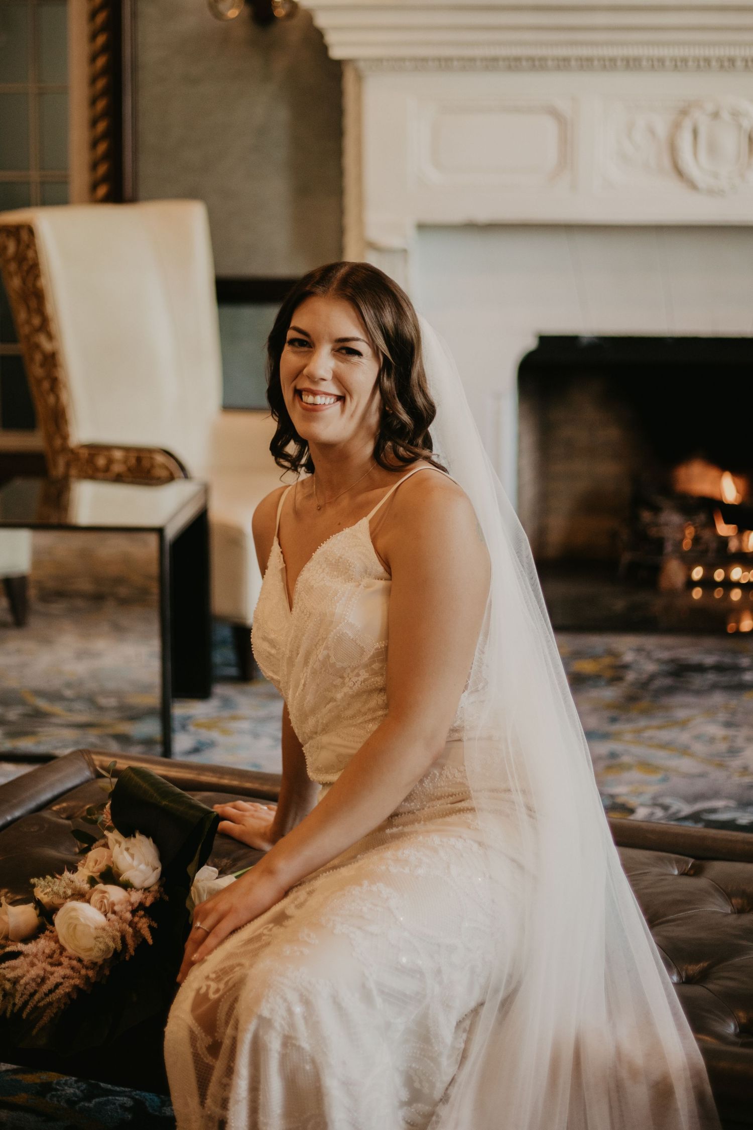 Bride sits elegantly by fireplace in vintage-style room wearing lace wedding gown and veil.