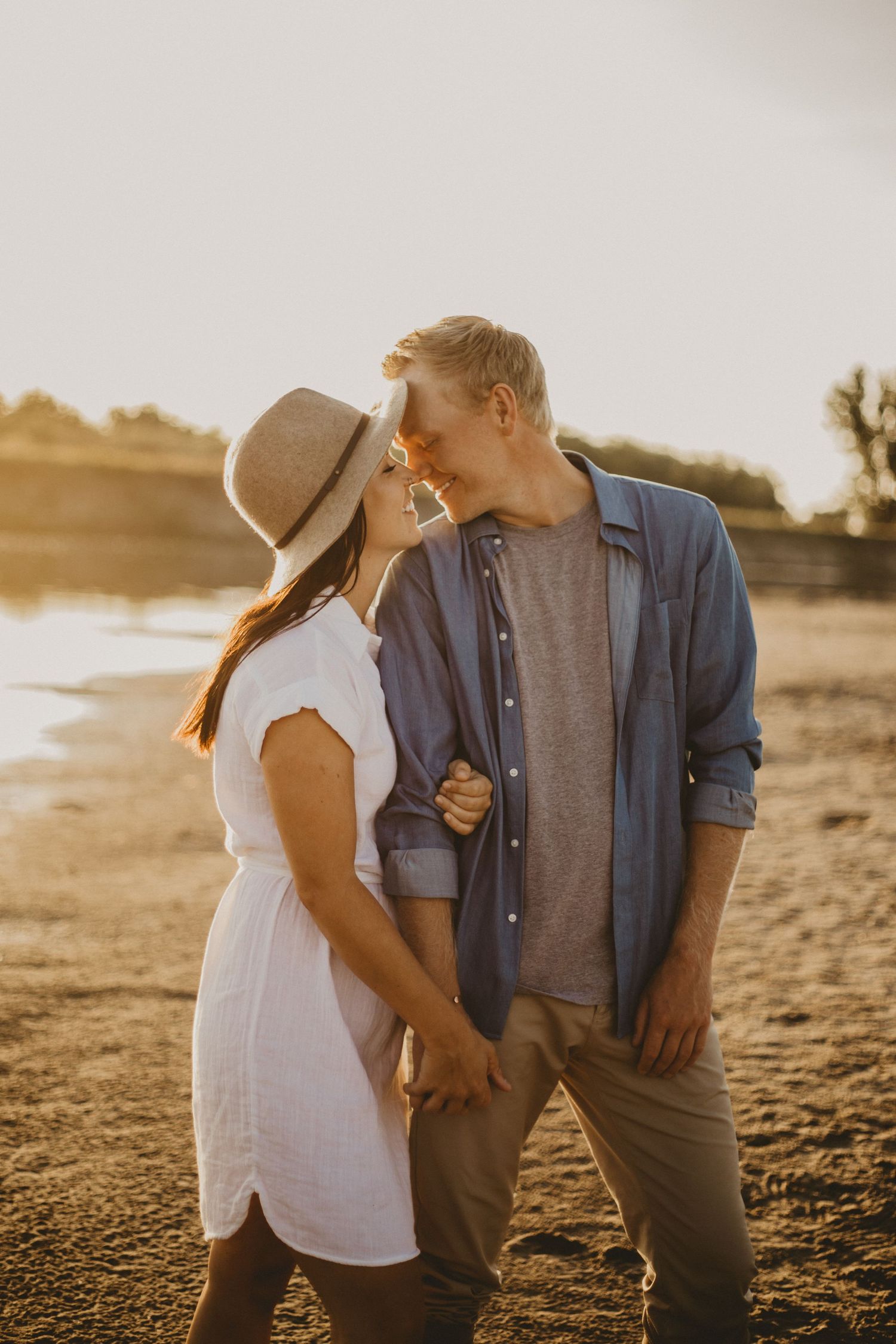 A tender moment between a couple on the beach at sunset wearing white and blue casual attire.