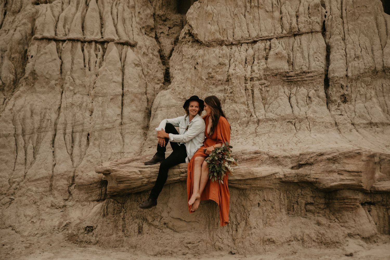 A couple sits together on a rocky ledge against textured clay cliffs during a romantic photoshoot.