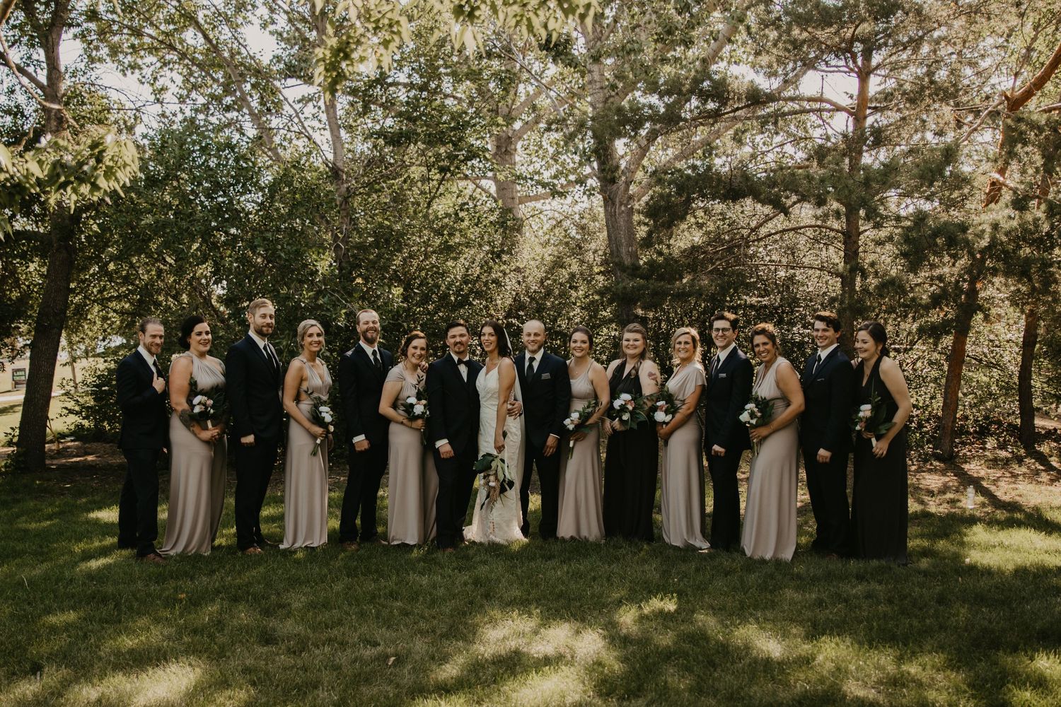 Large wedding party poses formally on grass with bridesmaids in matching taupe dresses.