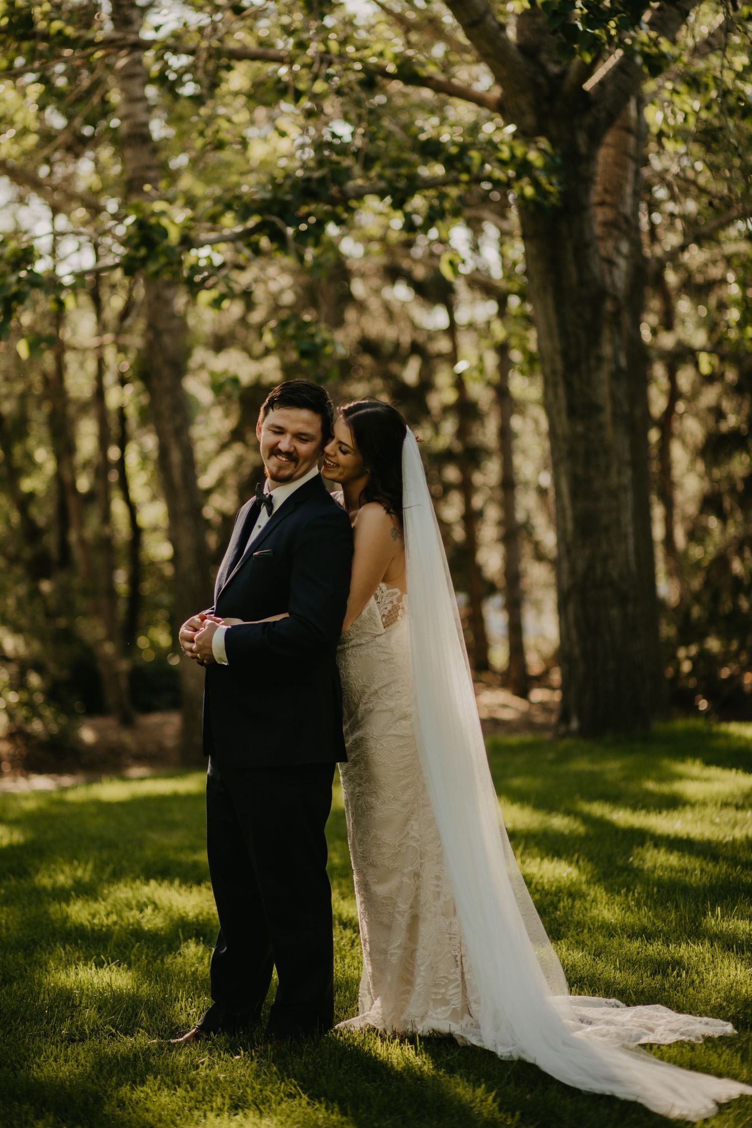 Joyful wedding couple poses in wooded garden setting with natural light filtering through trees.