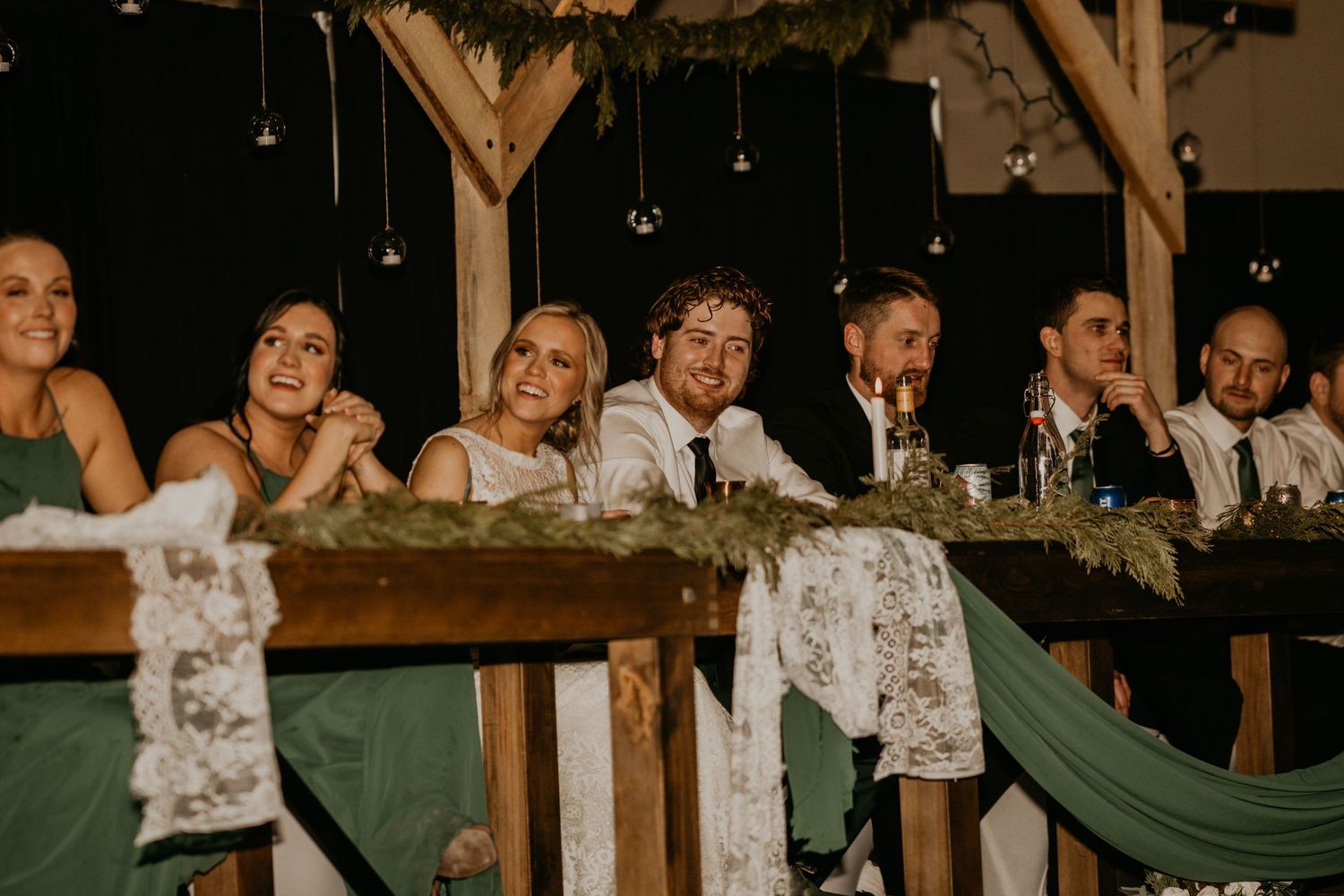 Wedding reception head table decorated with greenery and hanging lights in rustic venue.