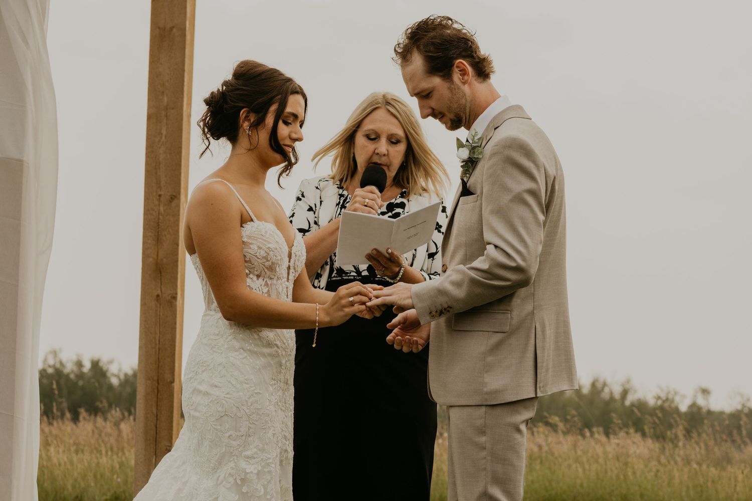 Wedding ceremony takes place under wooden arch with officiant and couple exchanging rings at outdoor venue.