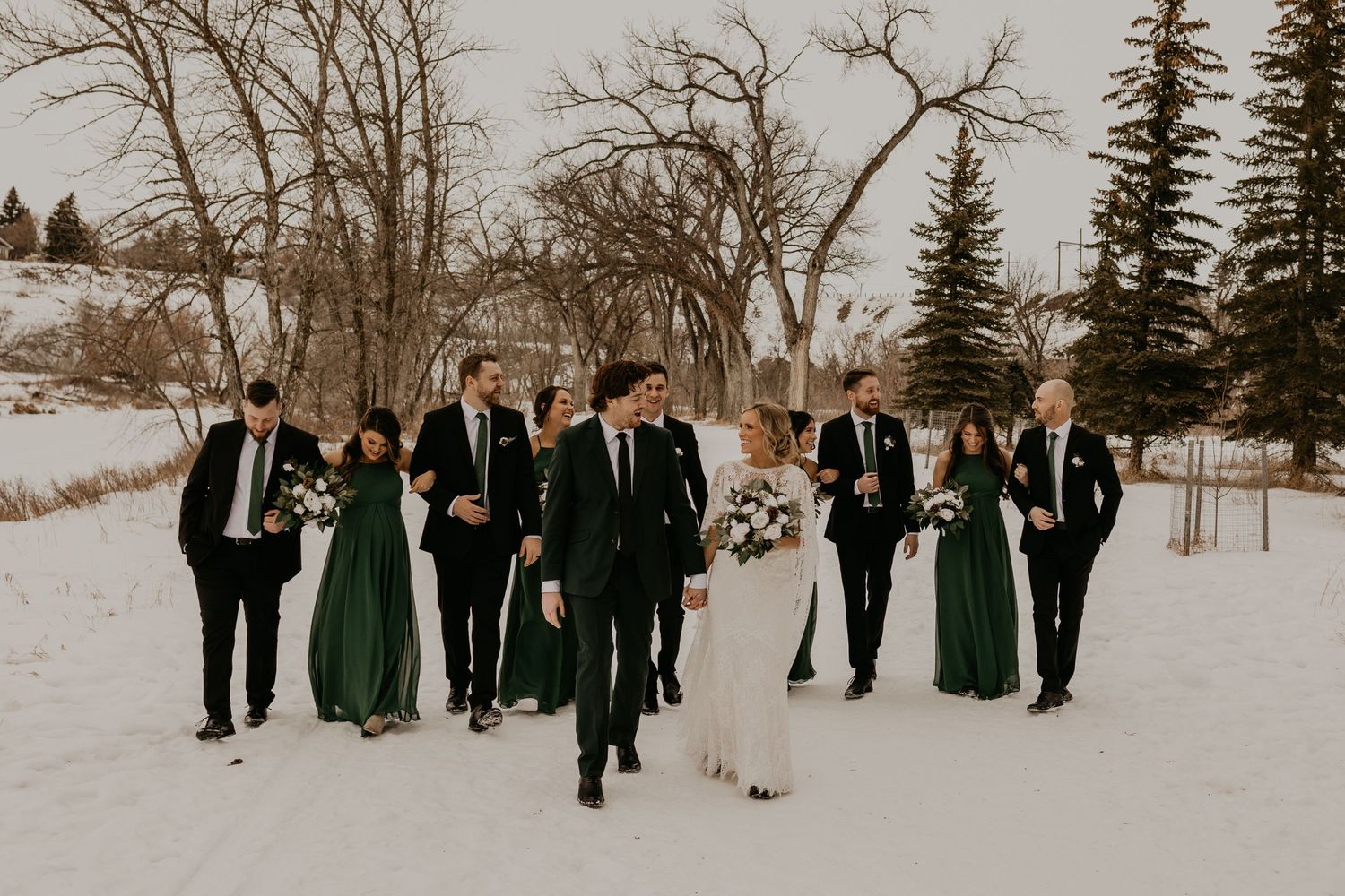Wedding party walking together through snowy winter landscape with evergreen trees and bridesmaids in emerald green dresses.