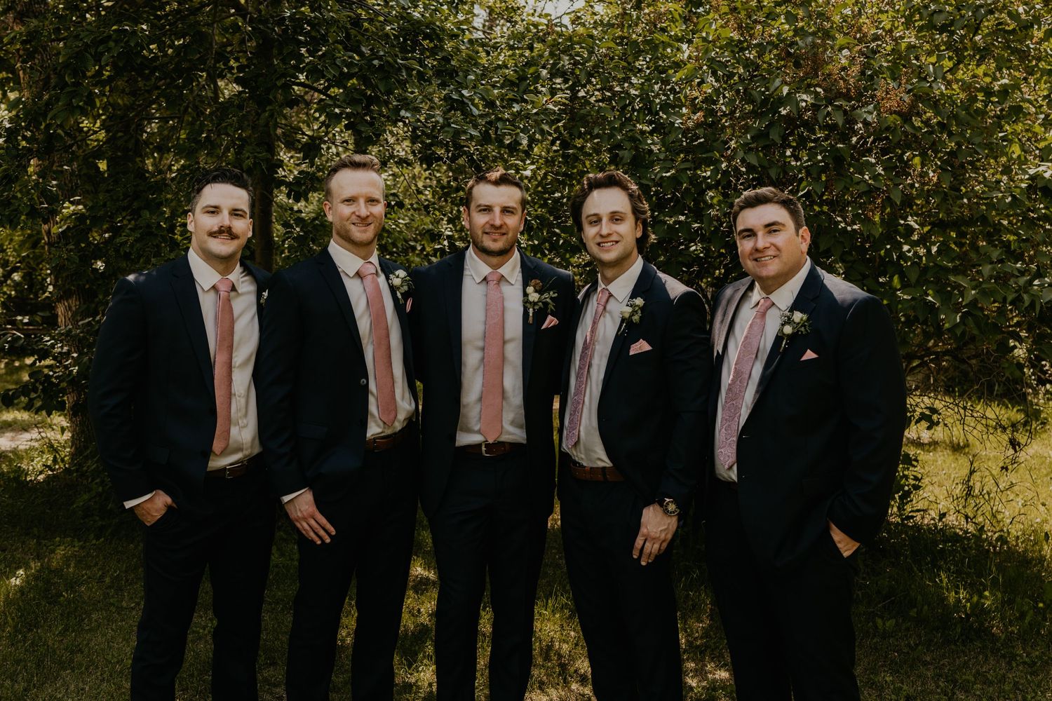 Groomsmen in dark suits with pink ties pose together against leafy backdrop.