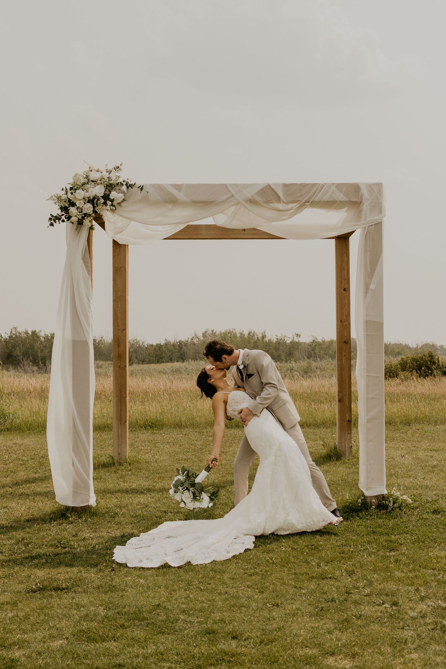 Romantic dip pose under white draped wedding arch in rustic outdoor setting.
