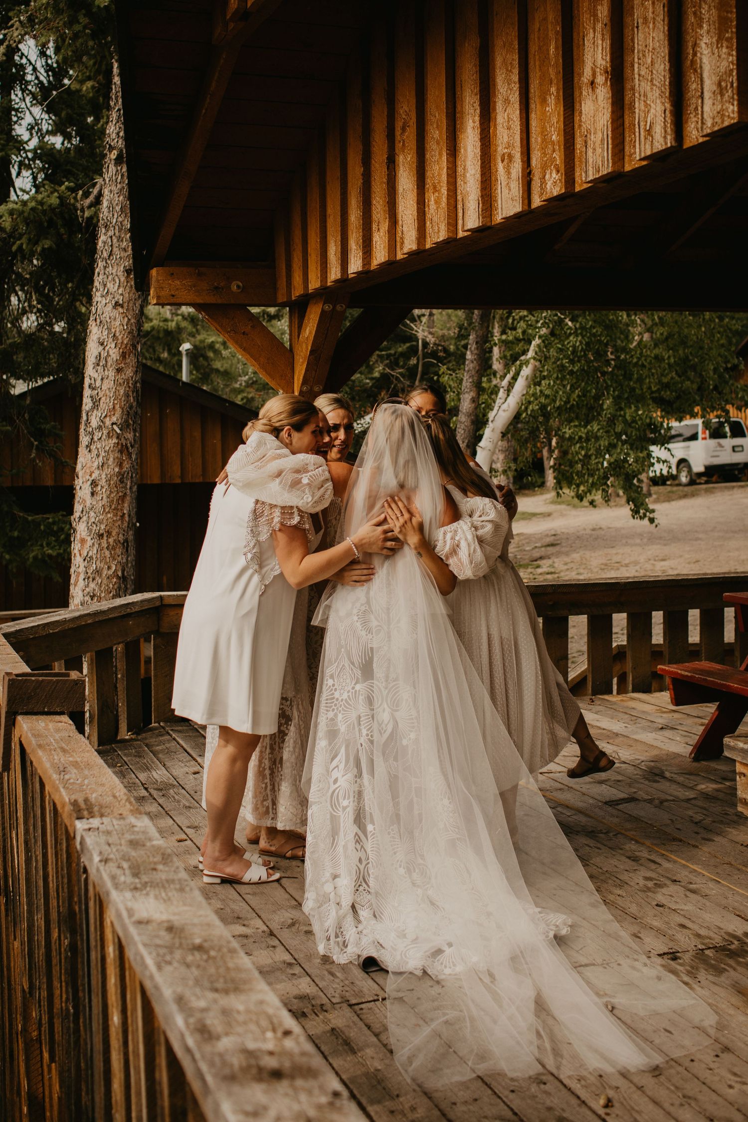 A bride in a flowing veil hugs her bridesmaids on a rustic wooden deck surrounded by trees.