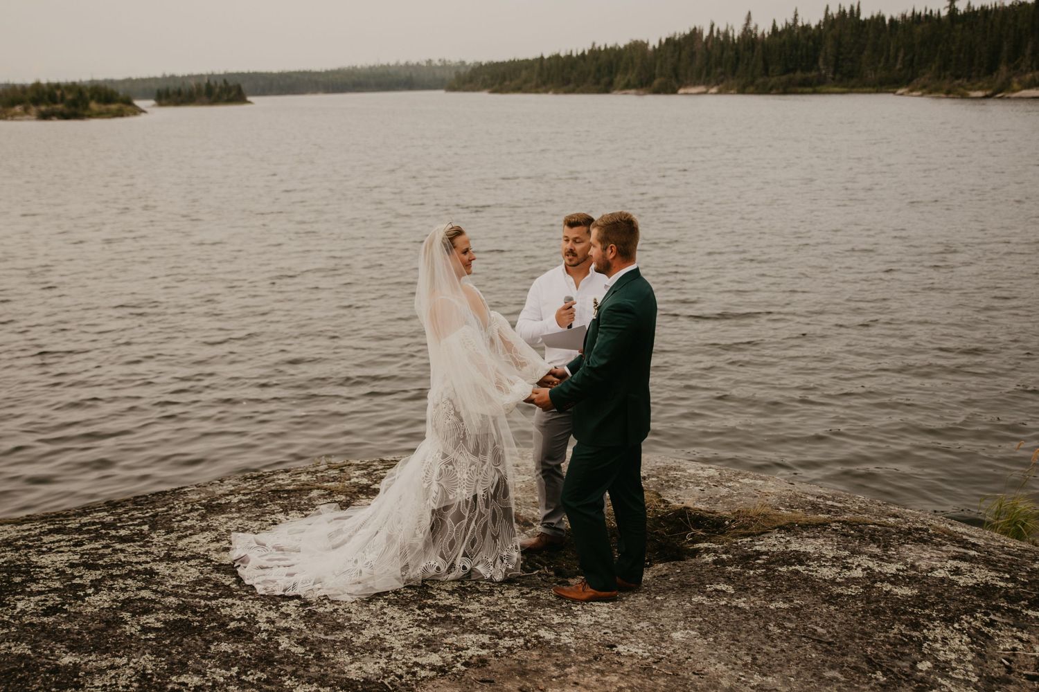 Wedding couple standing on rocky shore with lake and forest in background.