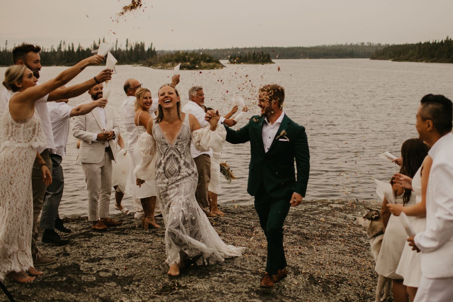 Wedding party celebrating and throwing confetti by a lake with evergreen forest backdrop.
