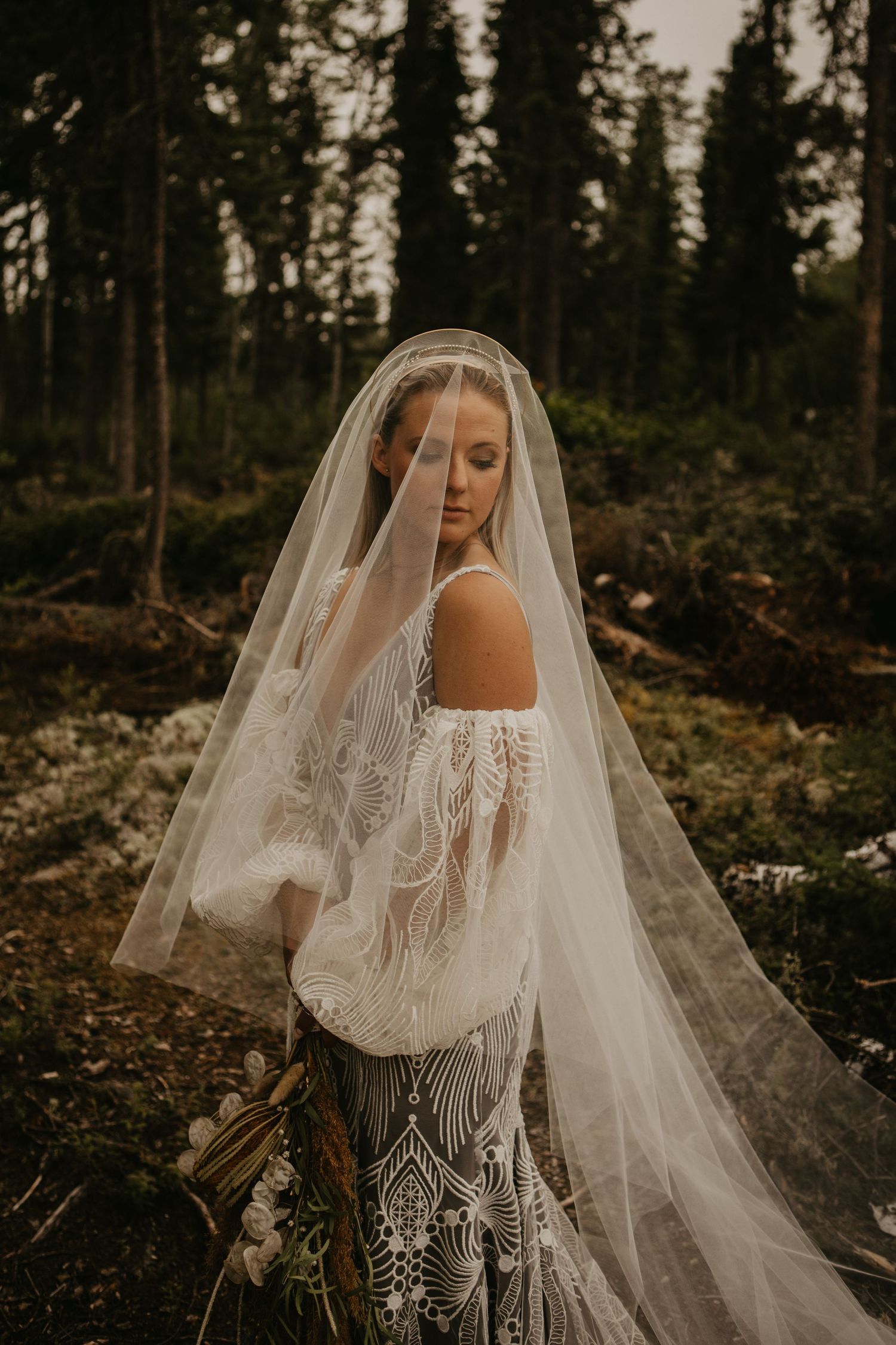 Bride in lace gown with flowing veil poses in moody forest setting at sunset.