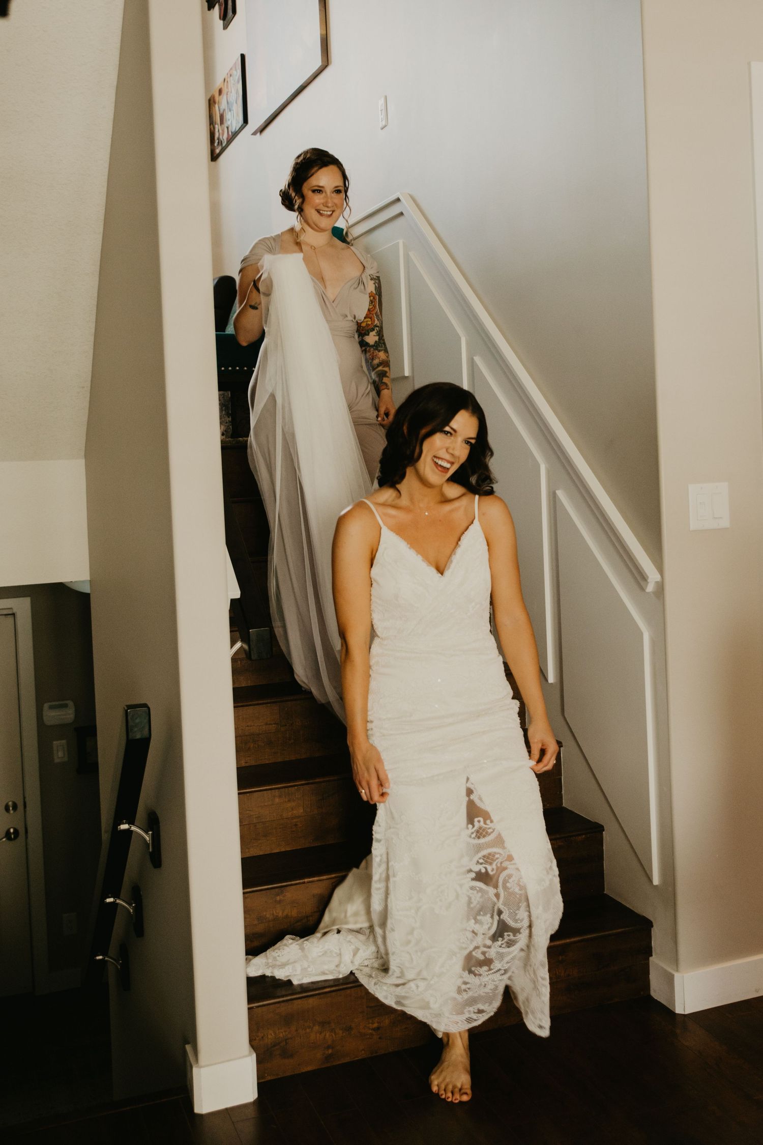 Two people in white wedding gowns descending wooden staircase.