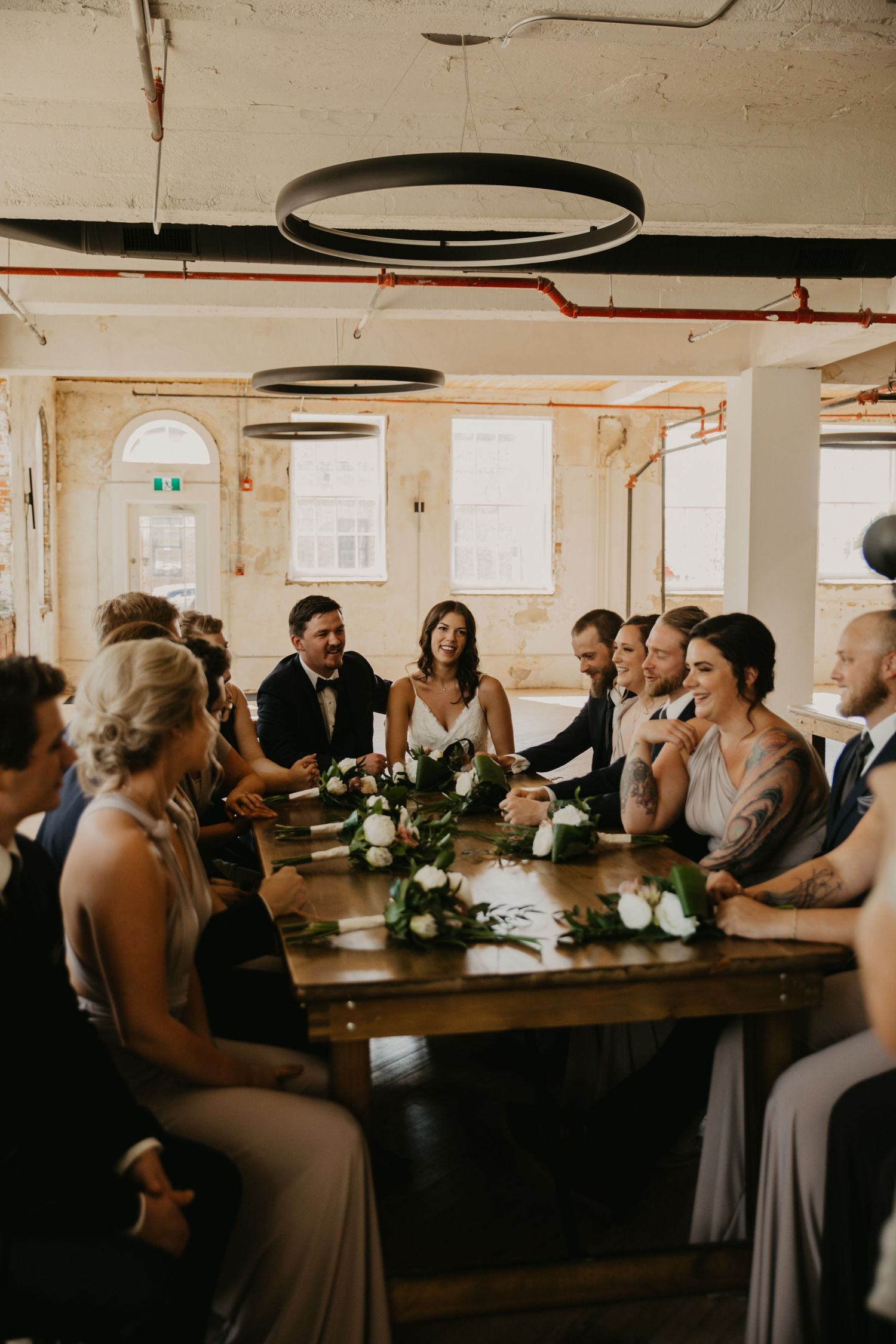 Wedding party gathered around a rustic wooden table in an industrial-style room.